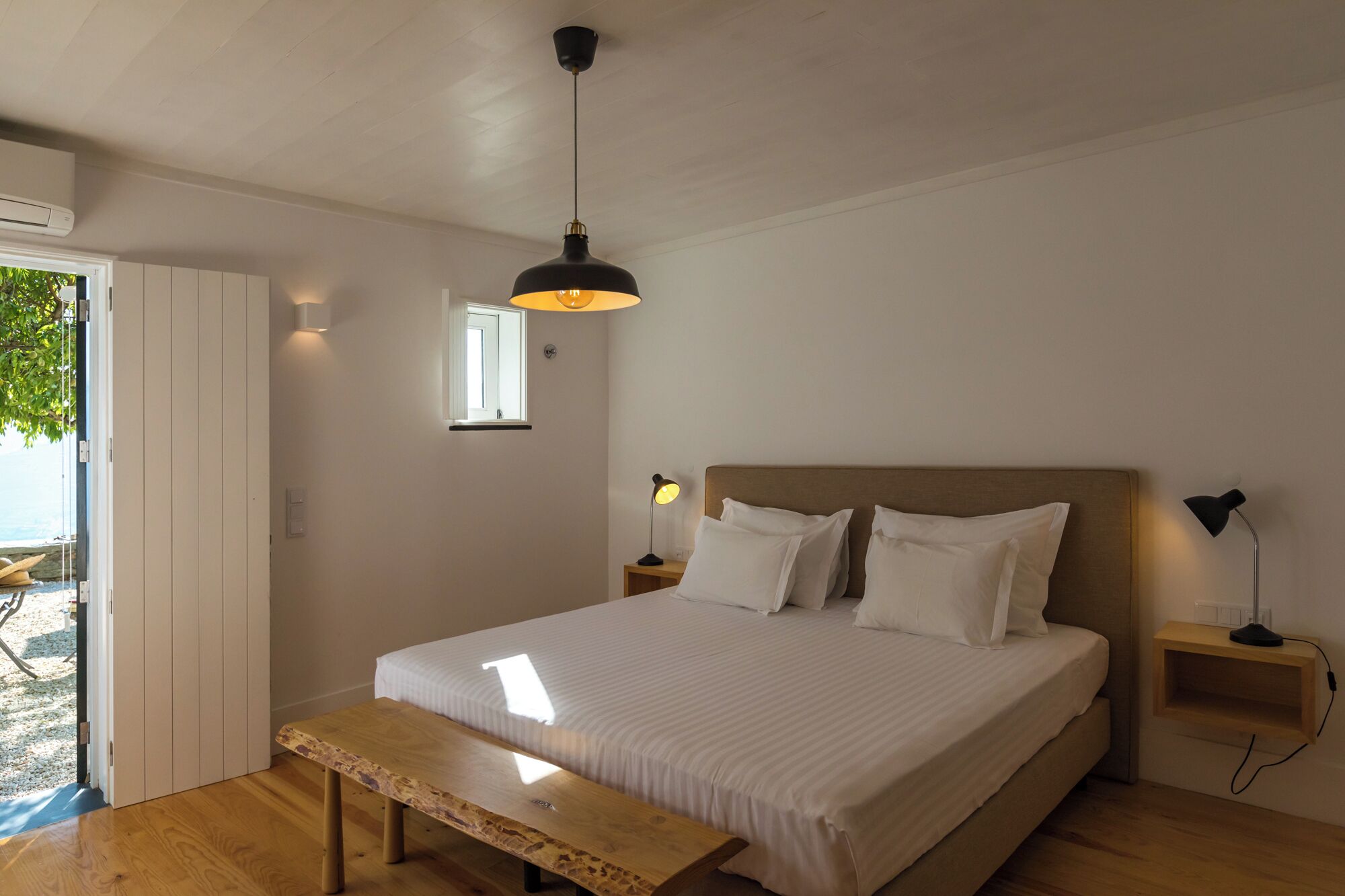 Un dormitorio con cama, mesitas de noche, un banco, una lámpara colgante y una ventana.