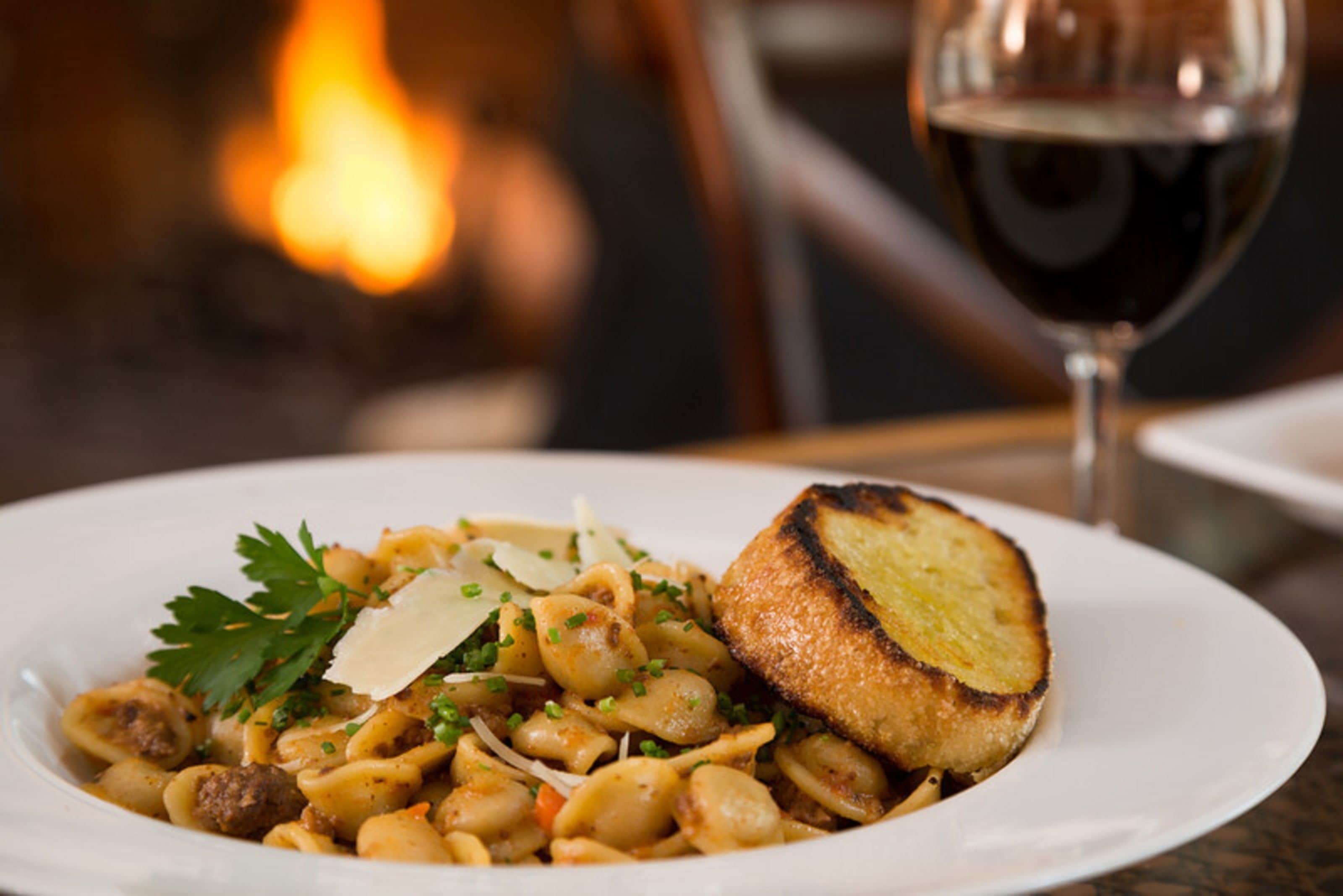 A plate of pasta with a toasted bread slice, garnished with parsley and grated cheese, accompanied by a glass of red wine.