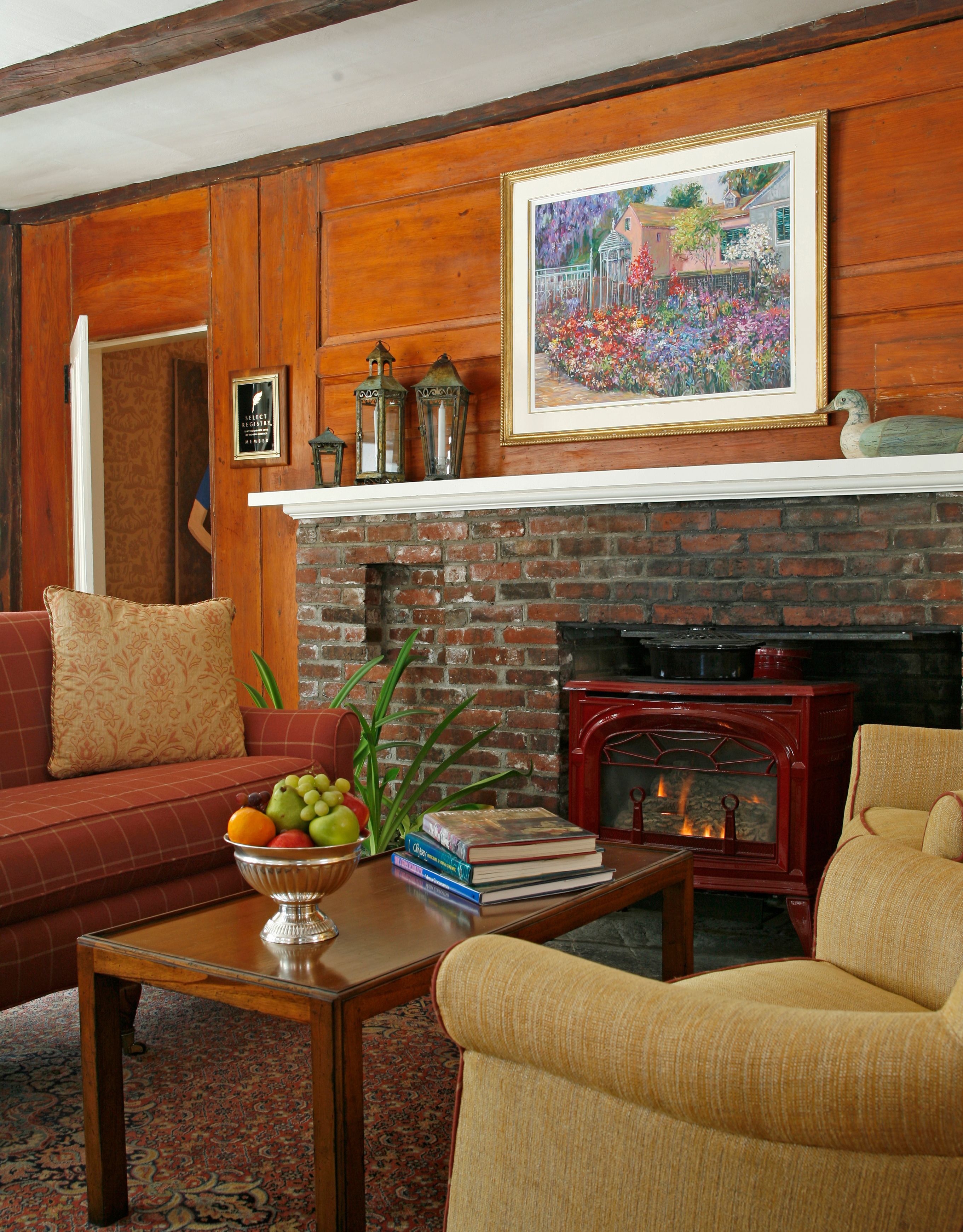 Una sala de estar acogedora con chimenea de ladrillo, paneles de madera, un sofá, dos sillones, una mesa de centro con frutas y libros, y un cuadro enmarcado sobre la chimenea.