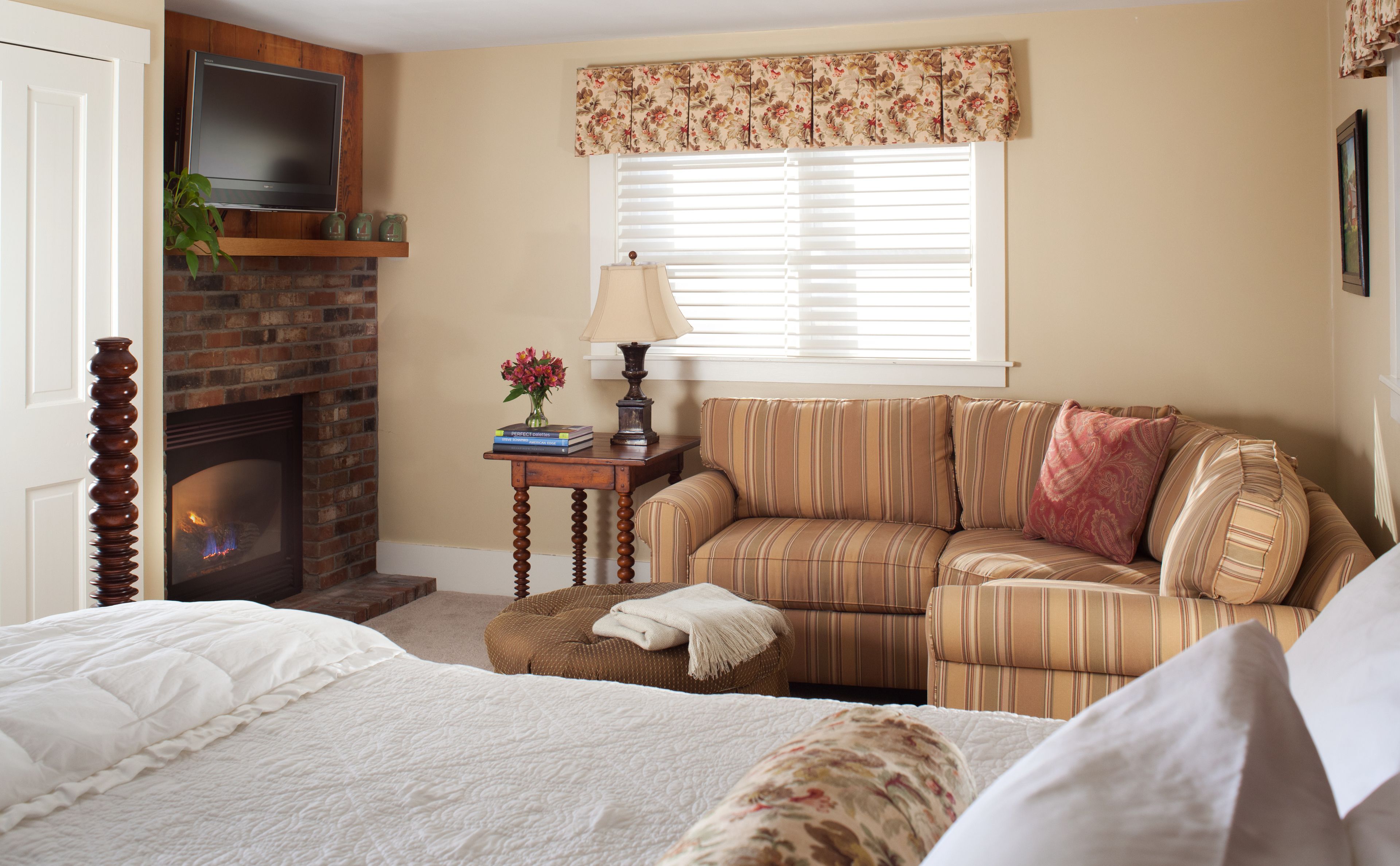 A cozy living room with a brick fireplace, a striped sofa, a small table with a lamp, and a bed with white bedding.