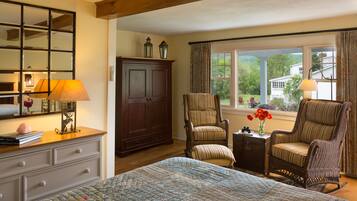 A bedroom with a large bed, a dresser, a wardrobe, two upholstered chairs, a small table with a lamp, and a window with curtains.