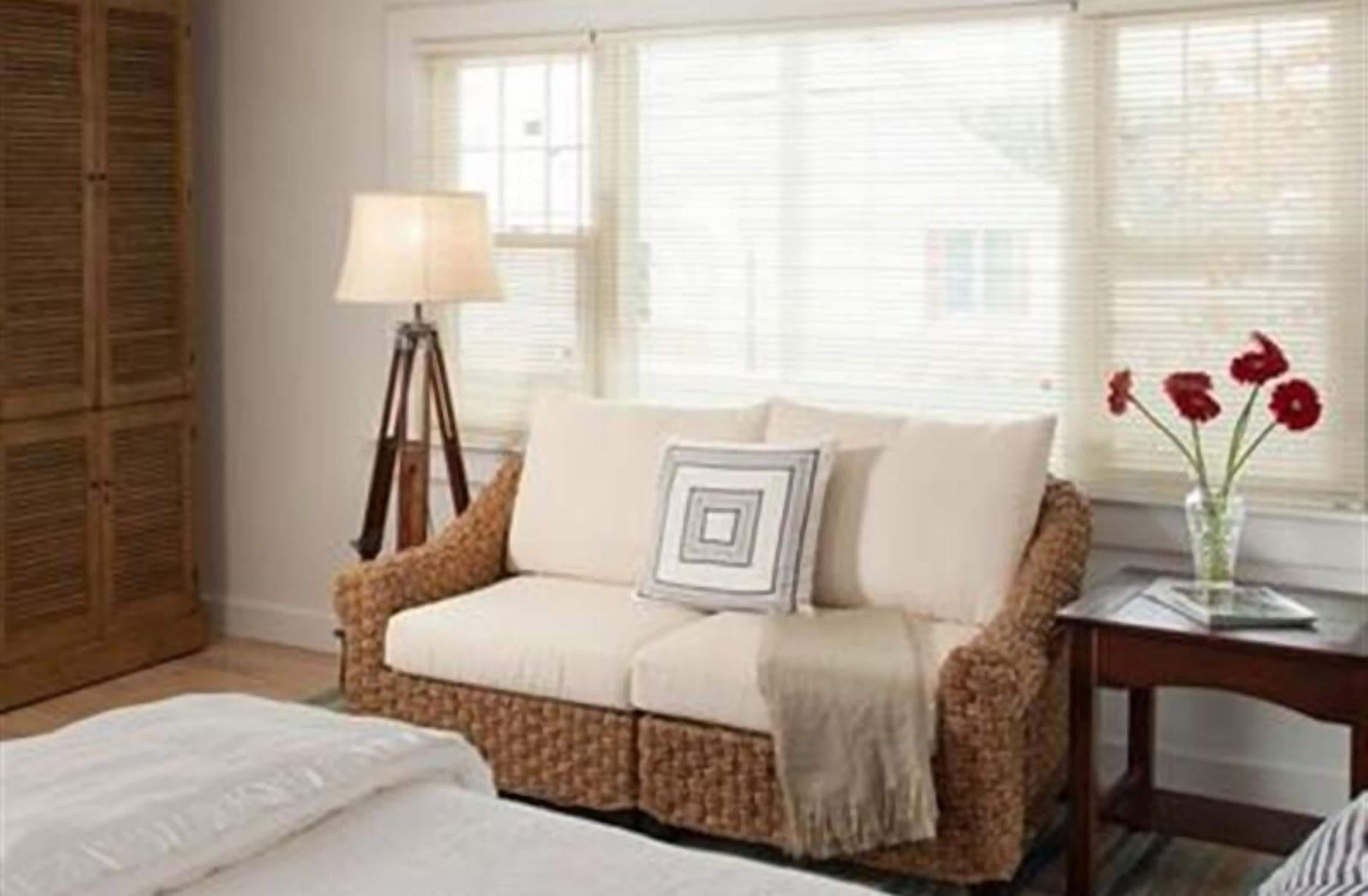 A room with a wicker sofa, a lamp, a vase with flowers, and a window with blinds.