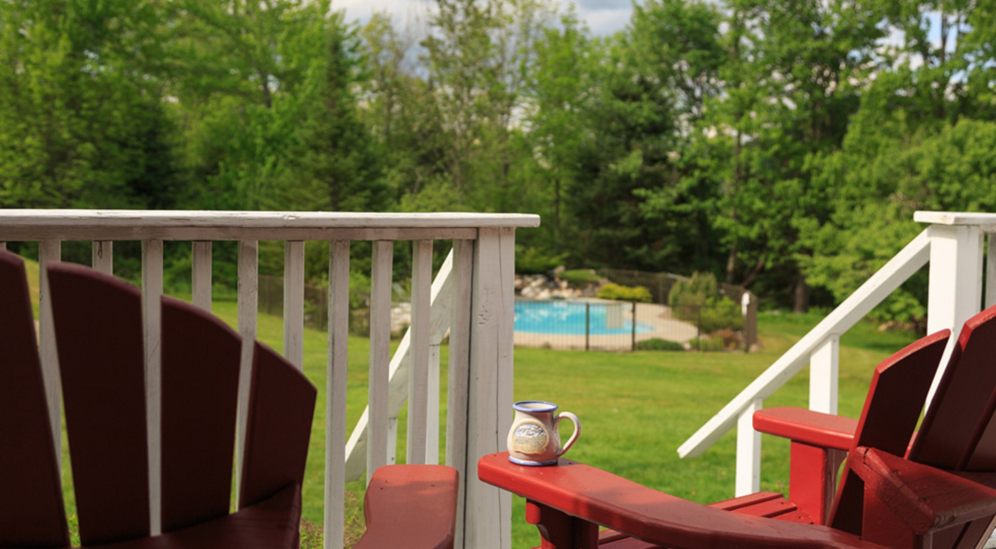 A deck with red chairs and a white railing overlooking a backyard with a pool and greenery.