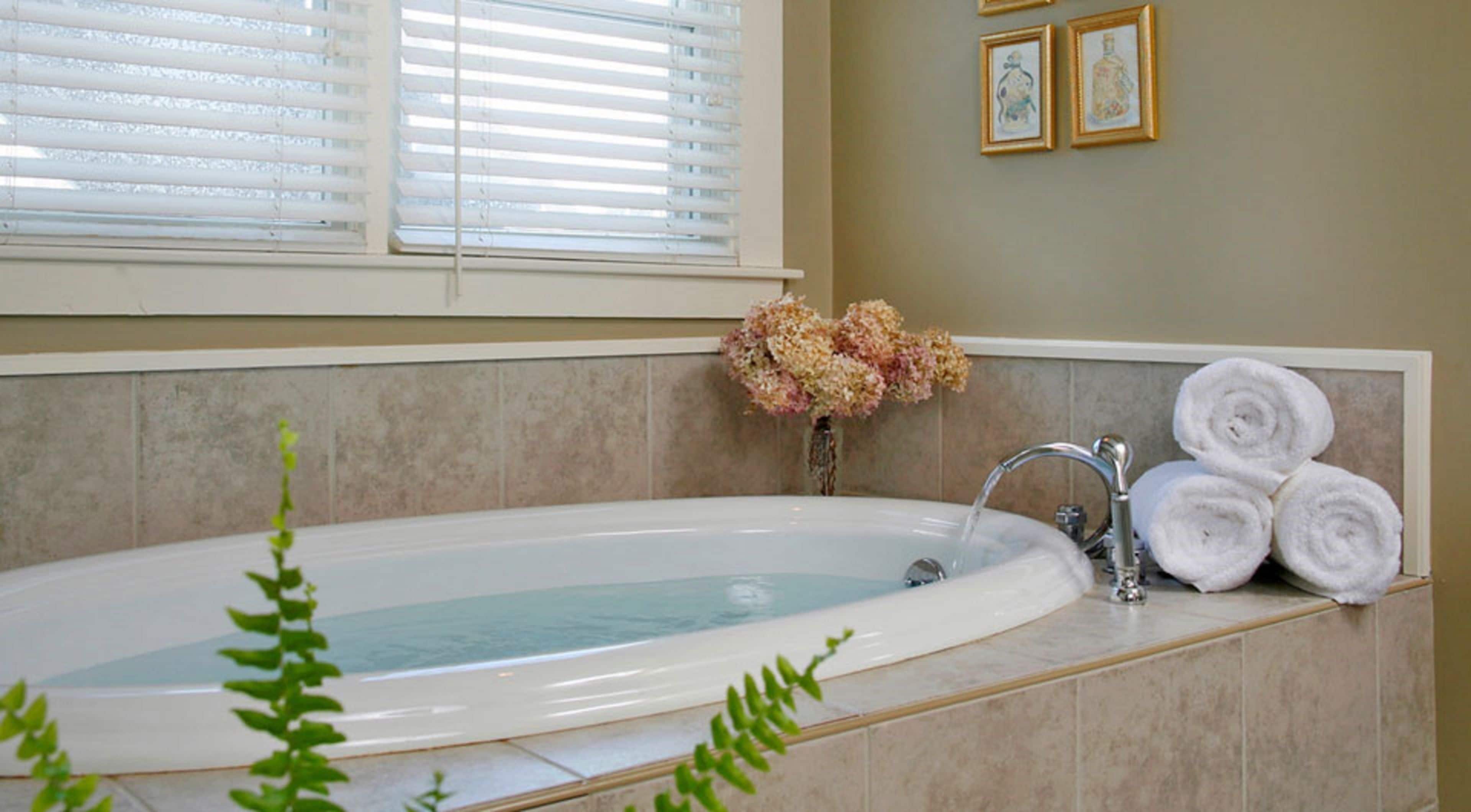 A bathroom with a bathtub, a sink, and a window with blinds. There are towels and a vase with flowers on the counter.