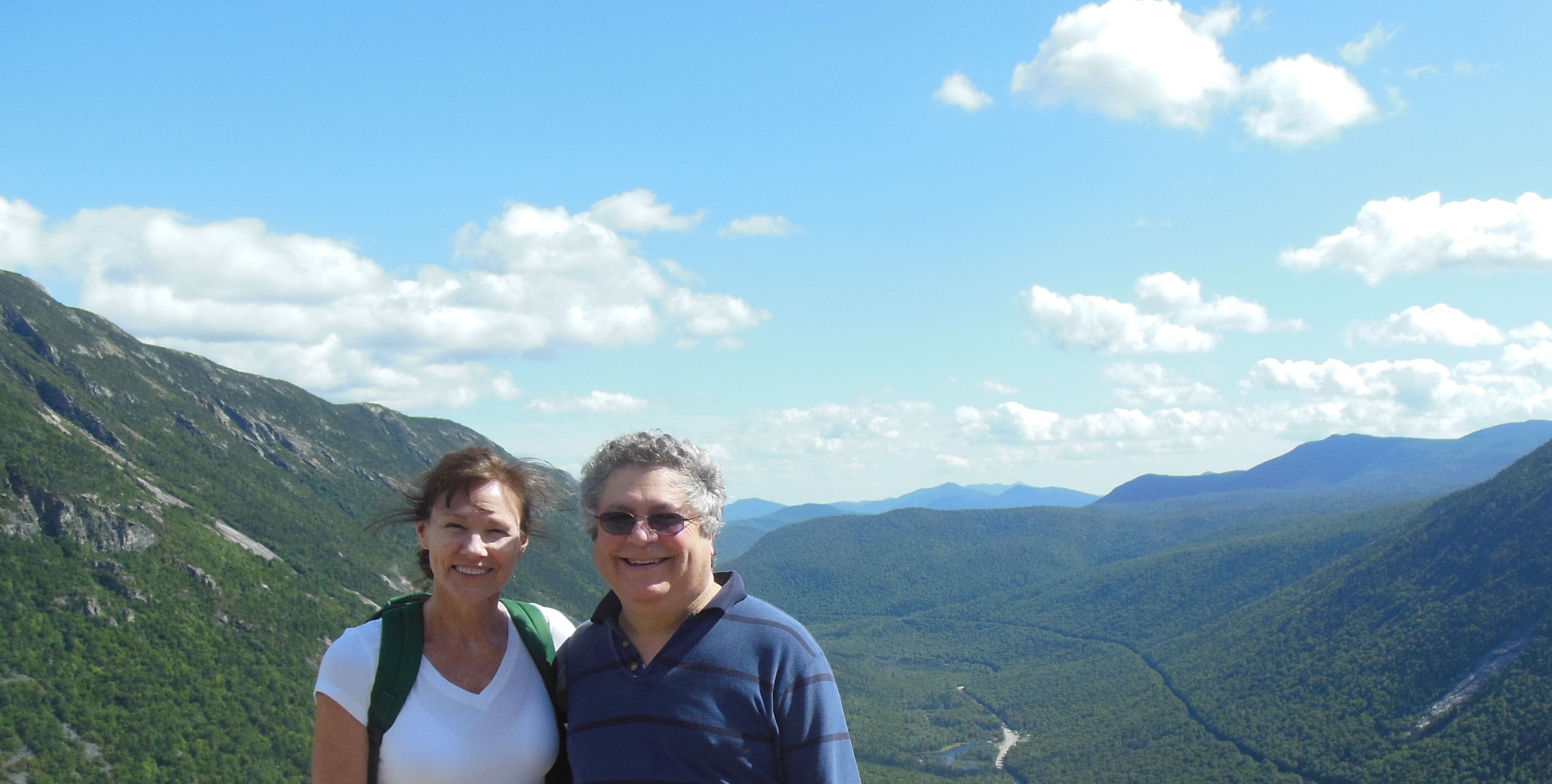 A couple standing on a mountain with a view of a valley and mountains in the background.