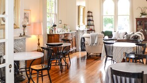 Une salle à manger spacieuse avec un plancher en bois, des murs blancs et un lustre. Plusieurs tables et chaises sont disposées pour accueillir les invités.