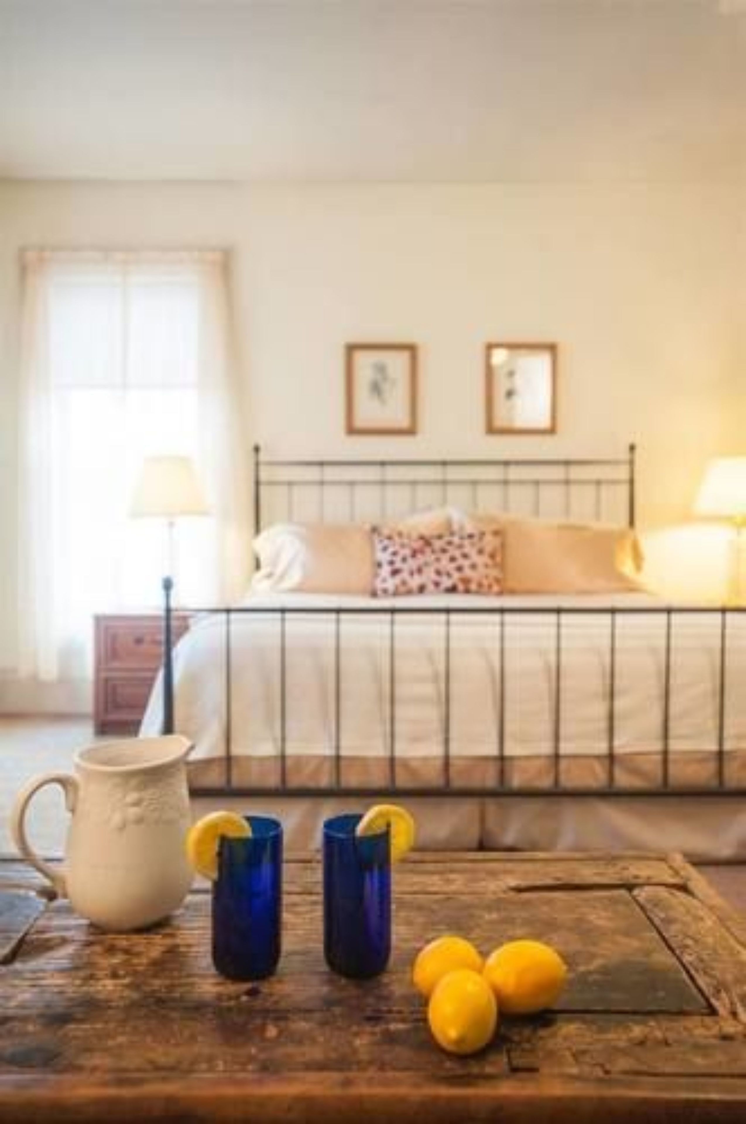 Un dormitorio con cama, lámparas de noche, una mesita de noche de madera y una mesa rústica con una jarra y vasos.