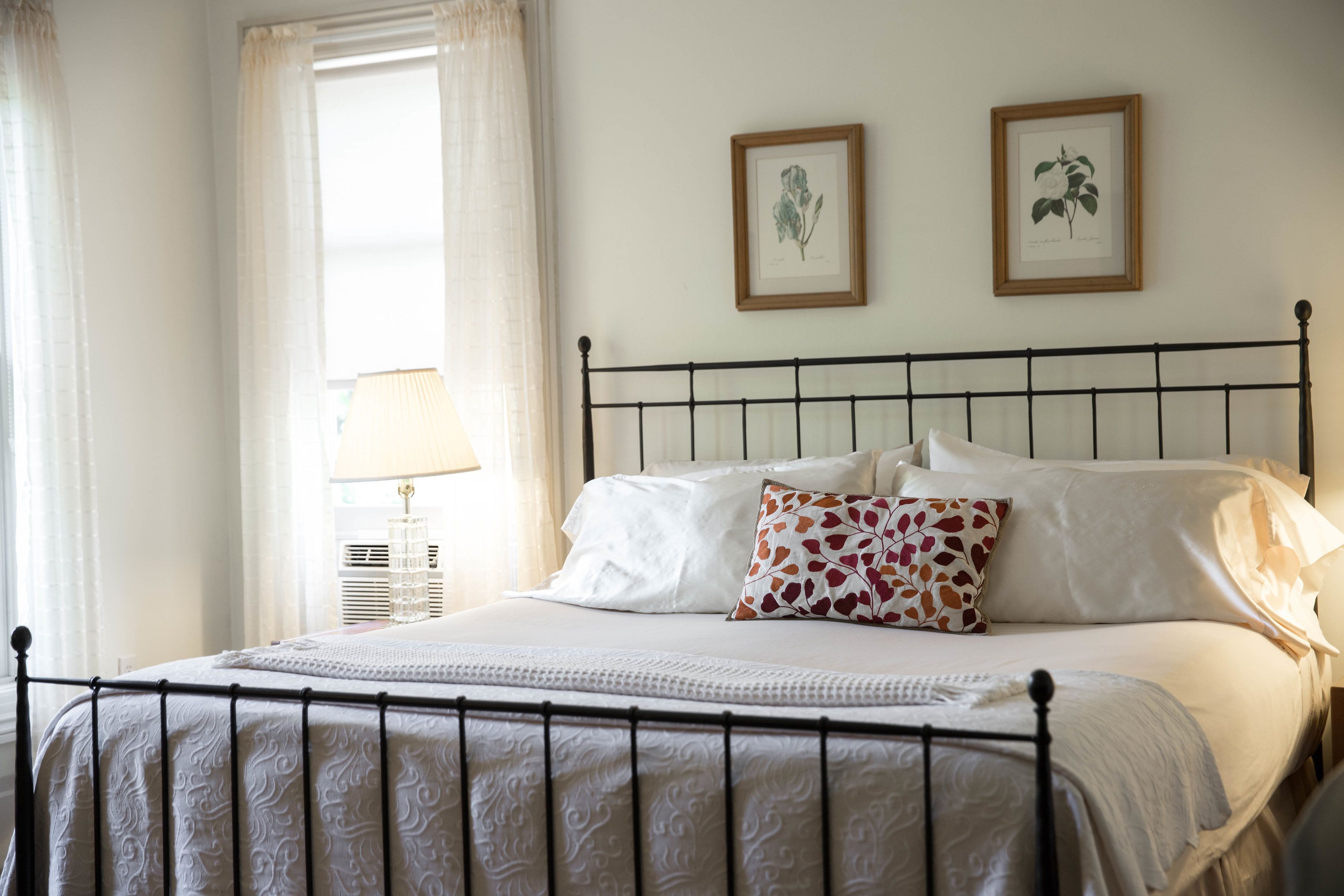 A neatly made bed with white linens and a patterned pillow, a bedside lamp, and two framed botanical prints on the wall.