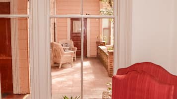 A room with a red armchair, a small table with a potted plant, and a view through a large window to an outdoor area with a brick wall, a wooden bench, and a hanging plant.