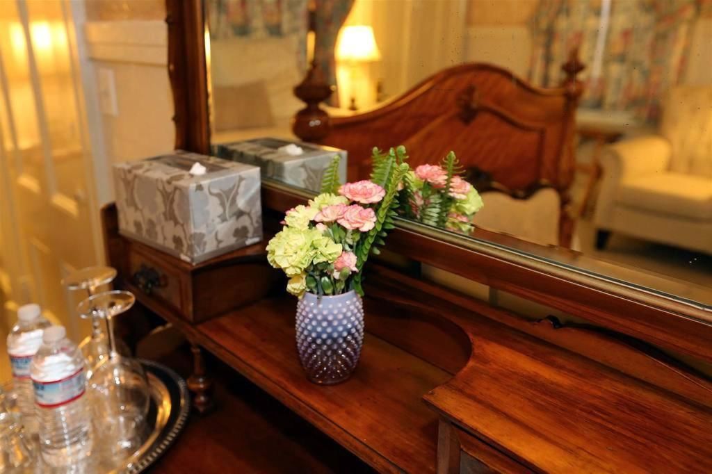 Une table de chevet en bois, ornée d’un vase de fleurs, d’un plateau avec des bouteilles d’eau et d’un miroir qui reflète un lit et une lampe.