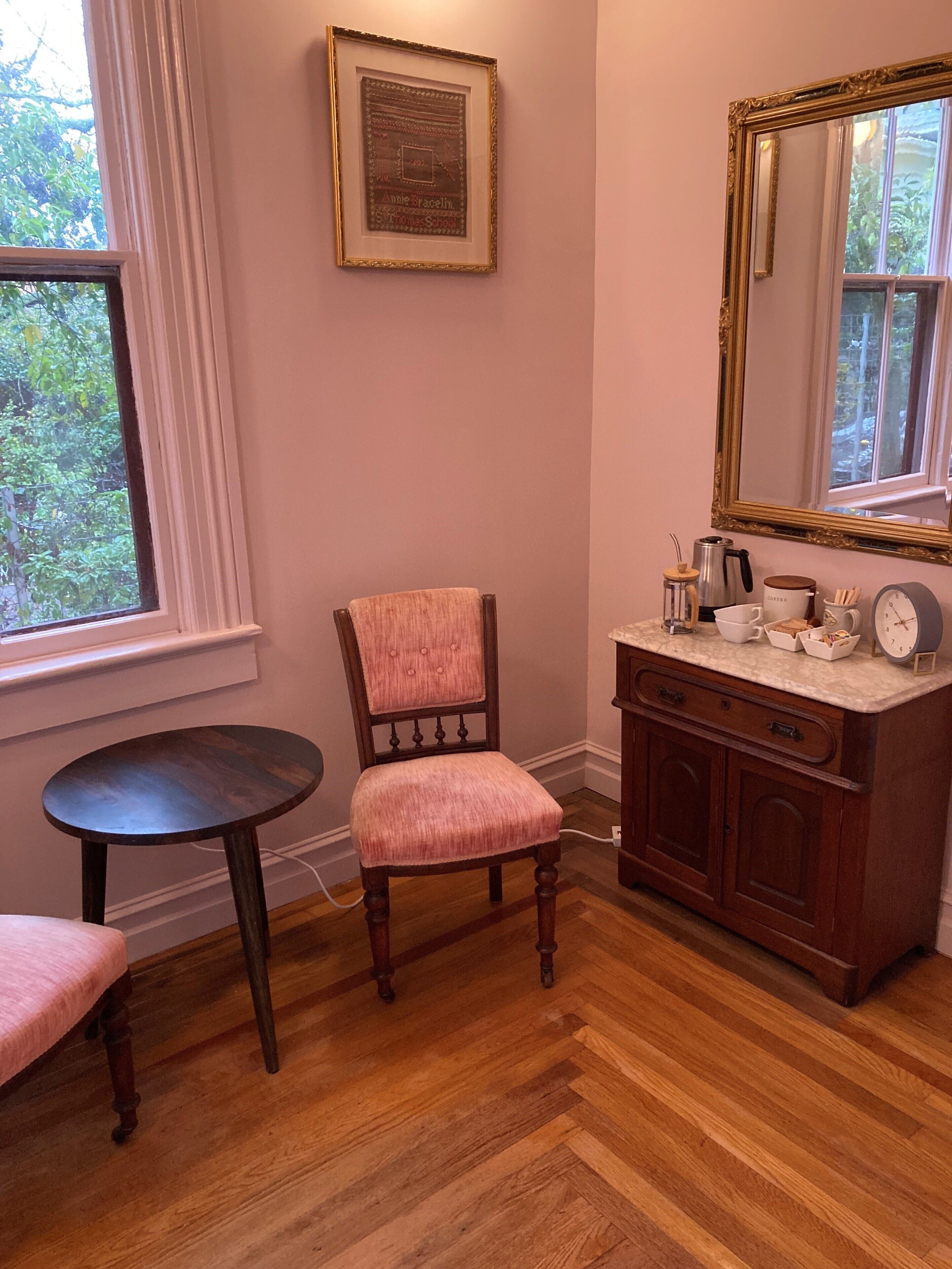 A room with a wooden herringbone floor, a wooden chair with a pink cushion, a small round table, a wooden cabinet with a mirror, and a framed picture on the wall.