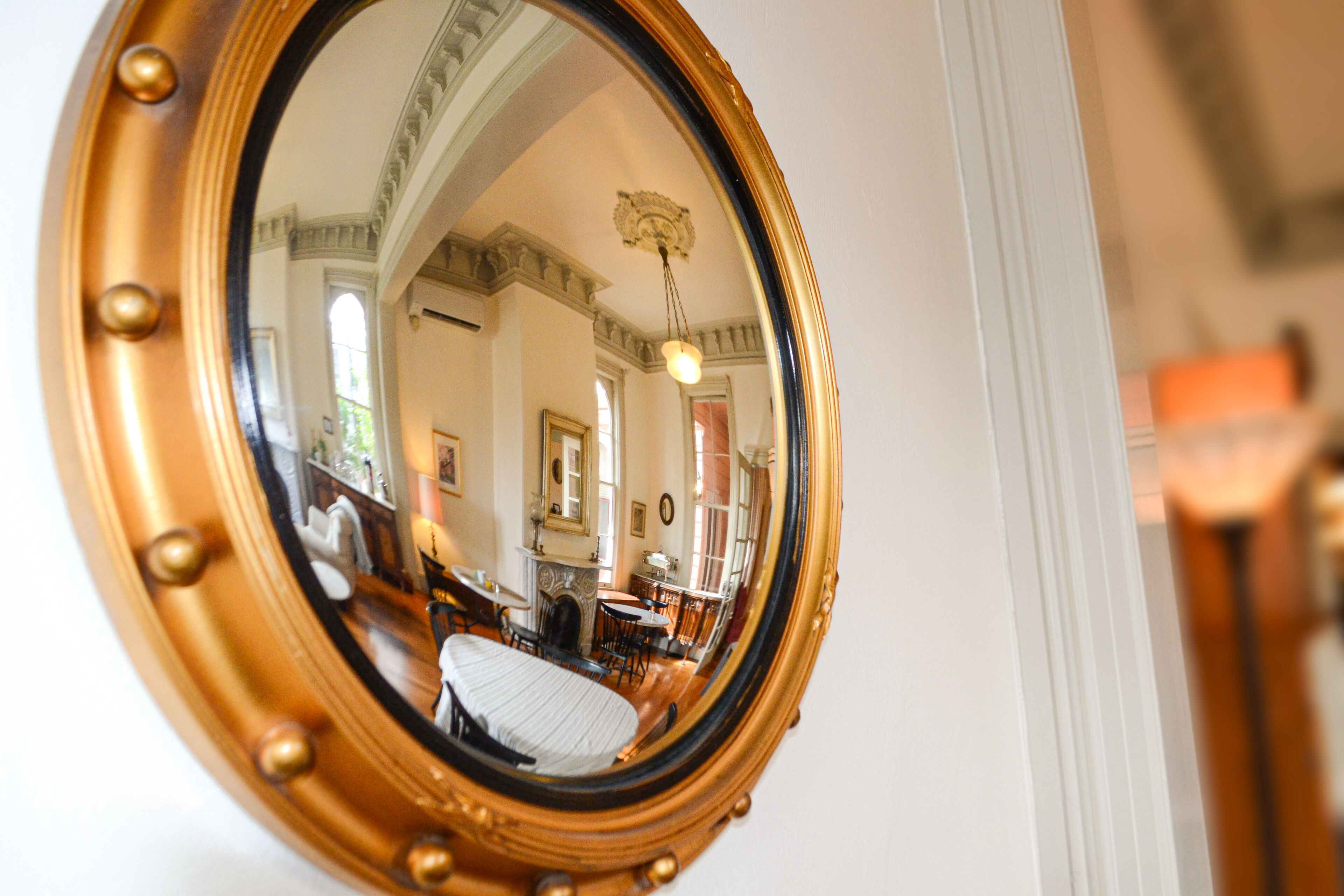 A round mirror with a gold frame reflecting a room with a fireplace, a chandelier, and a bed.