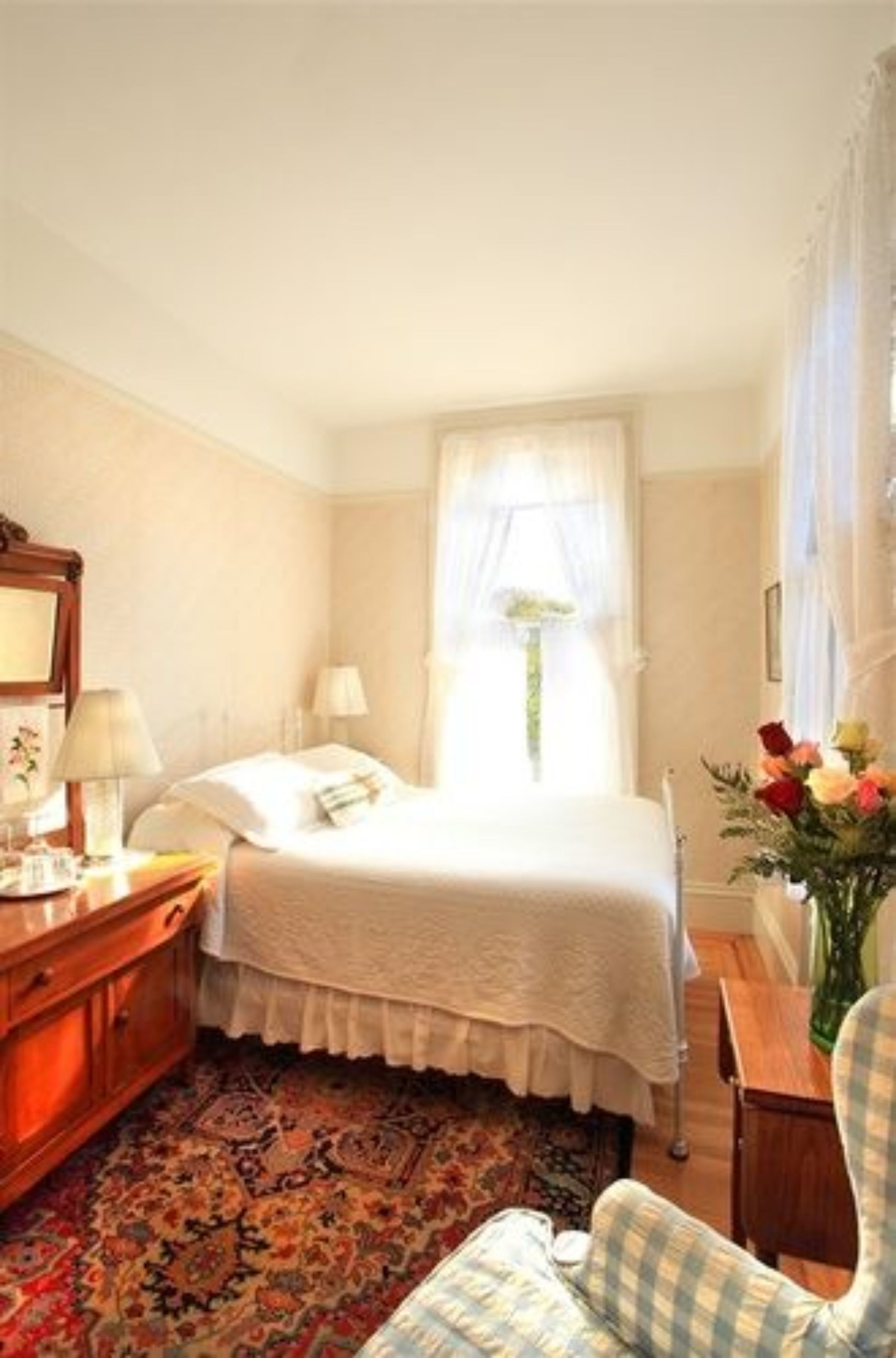 A hotel room with a bed, a desk with a lamp, a chair, a wooden dresser, and a window with curtains.