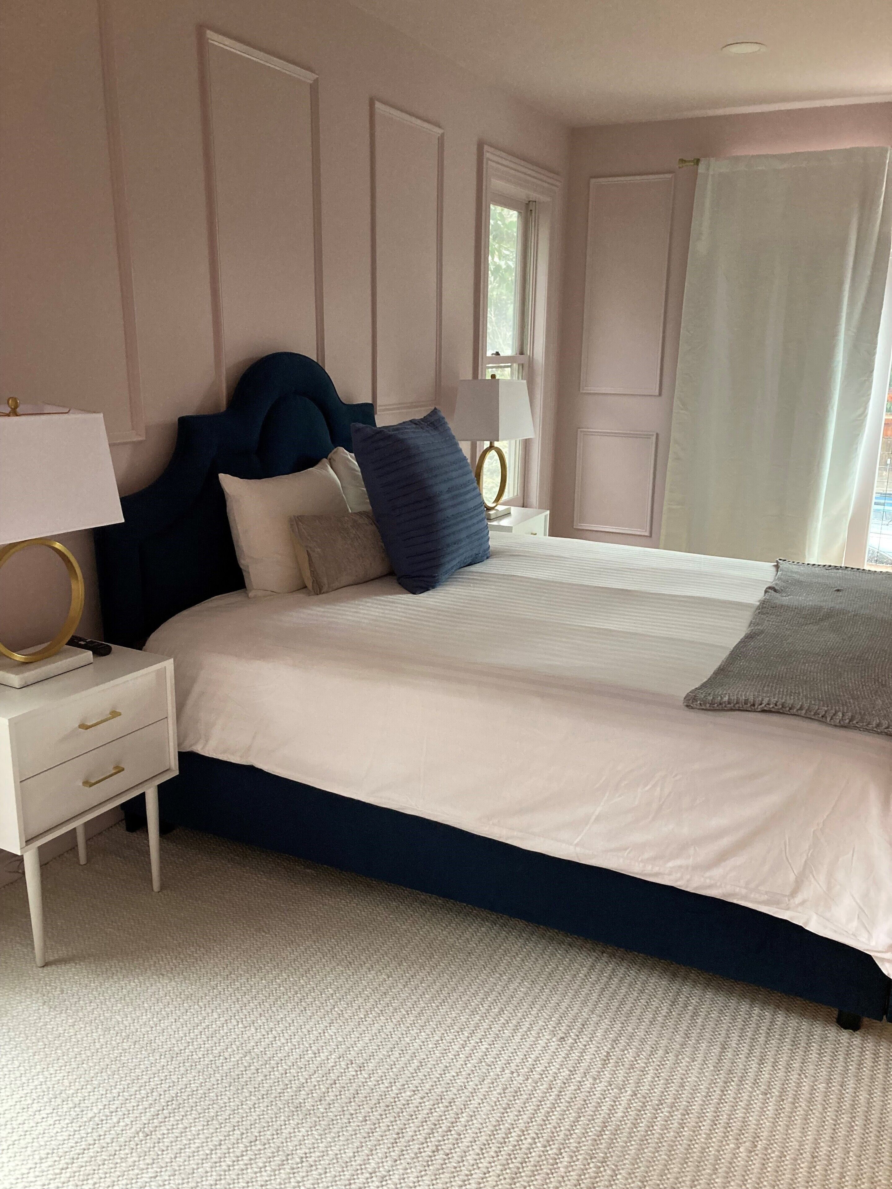 A neatly arranged bedroom with a large bed, a bedside table with a lamp, and a window with curtains.
