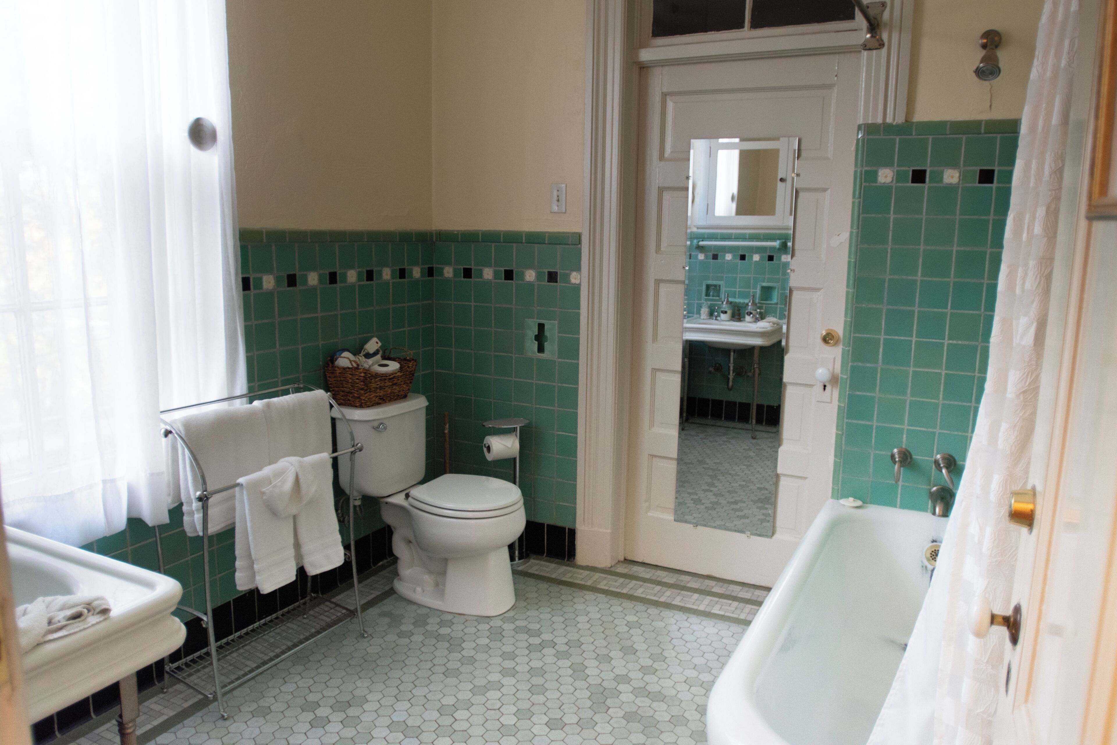 Een badkamer met toilet, wastafel en bad. Er is een handdoekhouder met handdoeken en een mandje op het toilet. De wanden zijn betegeld in groen en wit.