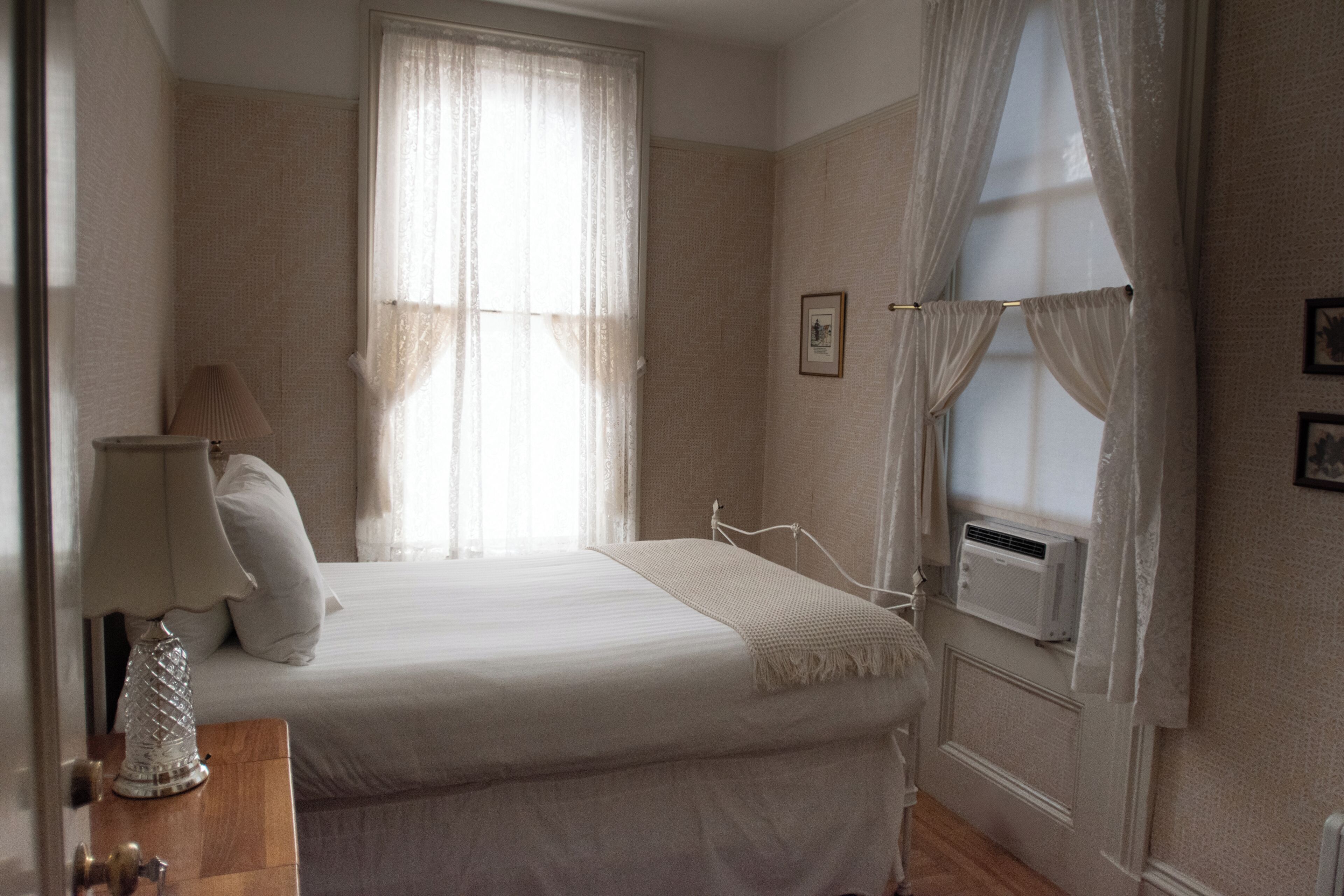 A bedroom with a bed, a bedside lamp, a window with curtains, and an air conditioning unit.