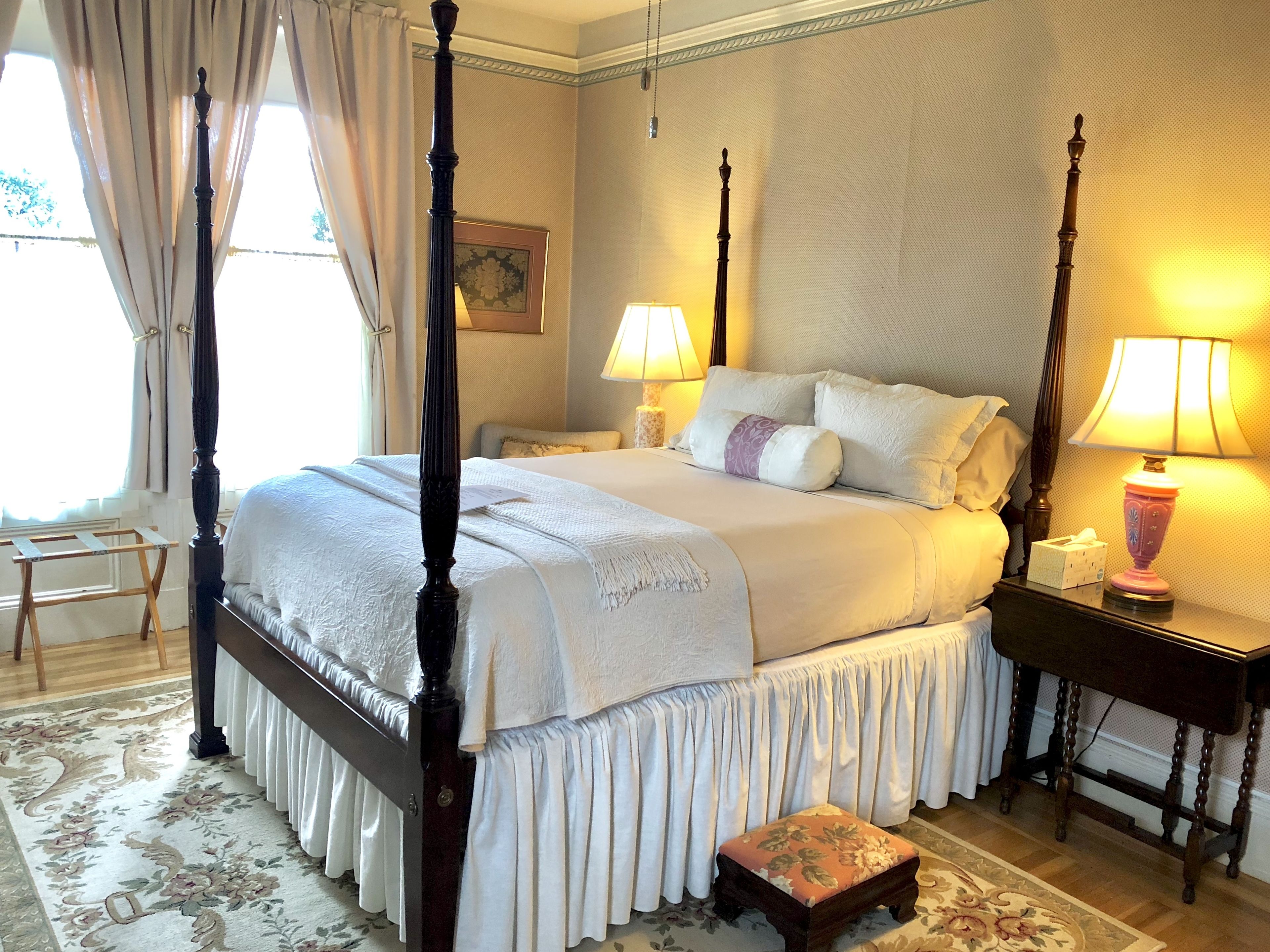 A four-poster bed with white bedding, two bedside lamps, and a floral-patterned ottoman.