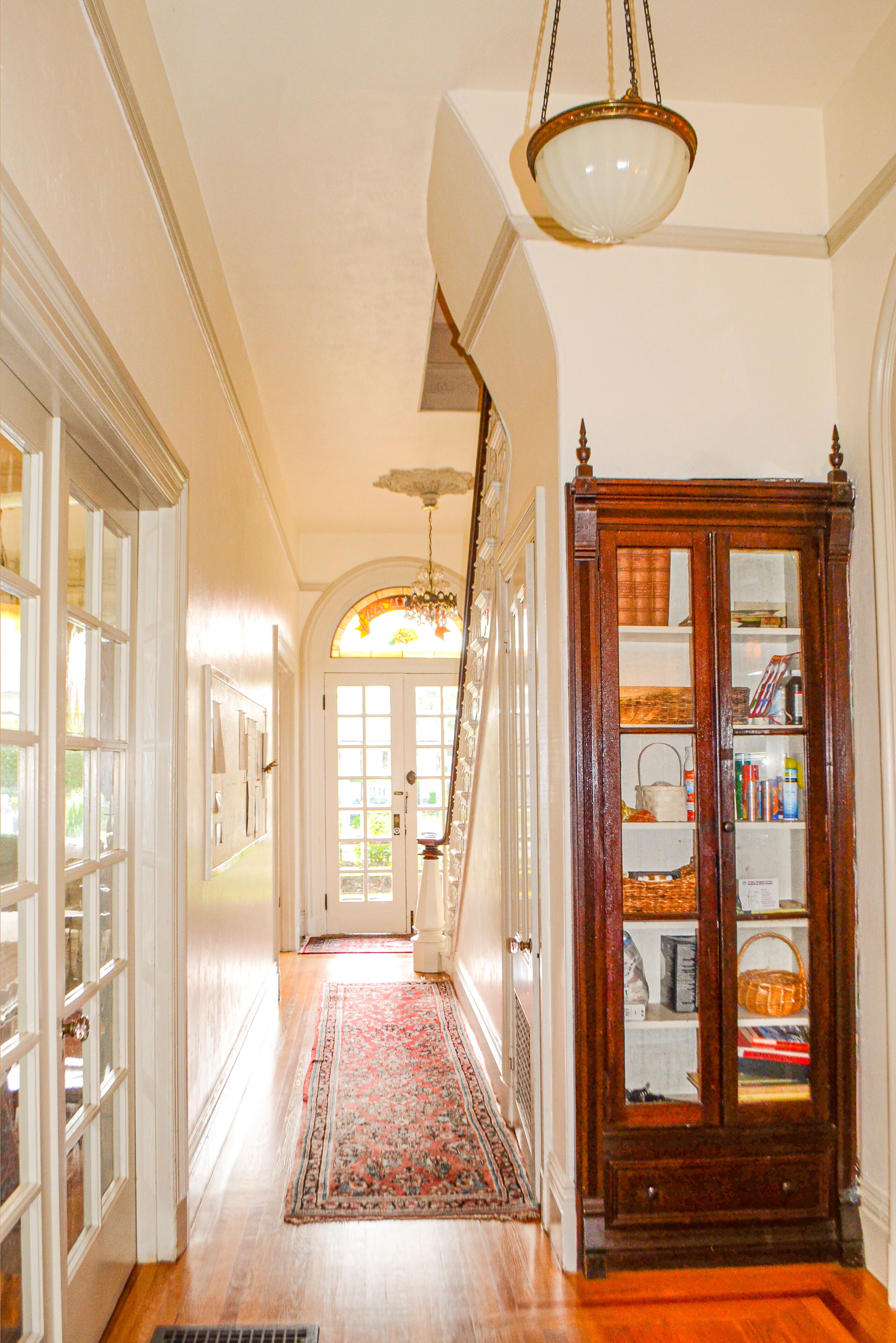 A hallway with a wooden floor, a patterned rug, a glass-fronted cabinet with shelves, and a chandelier.