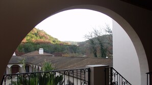 A balcony with a wrought iron railing, a view of a house with a tiled roof, and a landscape with trees and hills.