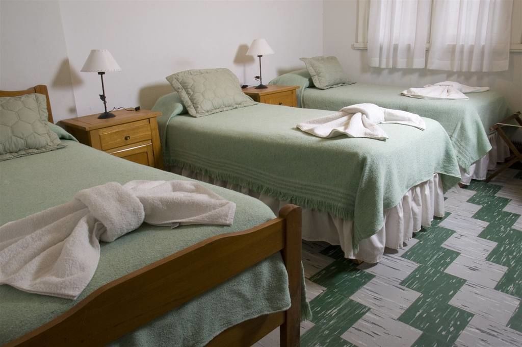 A room with two beds, each with a green bedspread and a white pillow. There are wooden nightstands with lamps, and a green and white checkered floor.