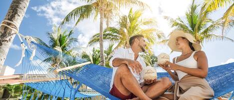 Un couple est assis dans un hamac, dans un cadre tropical, avec des palmiers et un ciel dégagé.
