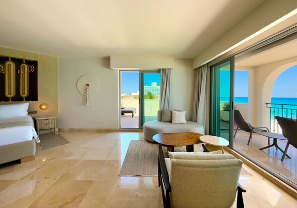 Habitación de hotel moderna con cama, zona de estar con mesa y sillas, y balcón con vistas al mar.
