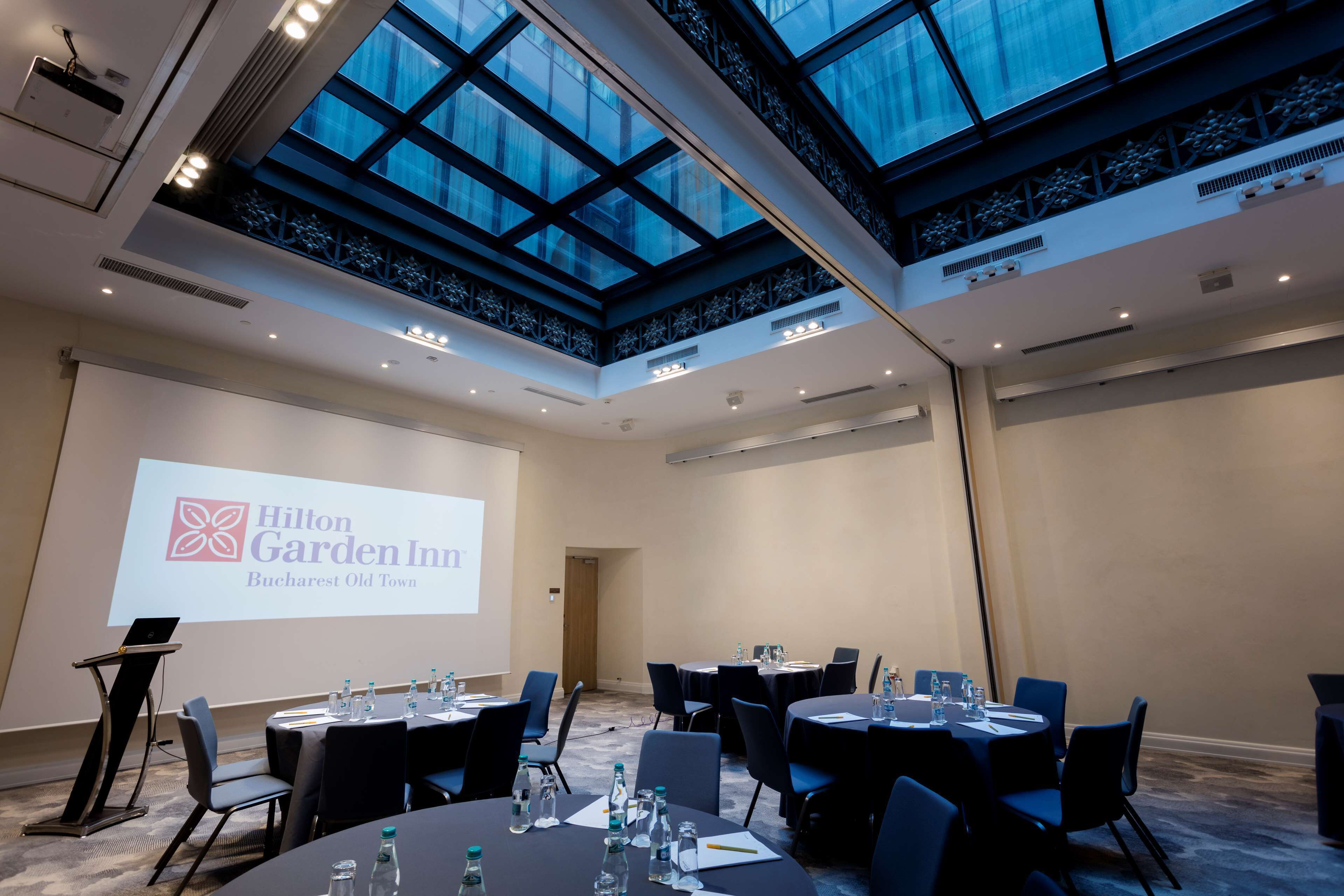 A conference room with a large screen displaying the Hilton Garden Inn logo, multiple tables with chairs, and a high ceiling with skylights.