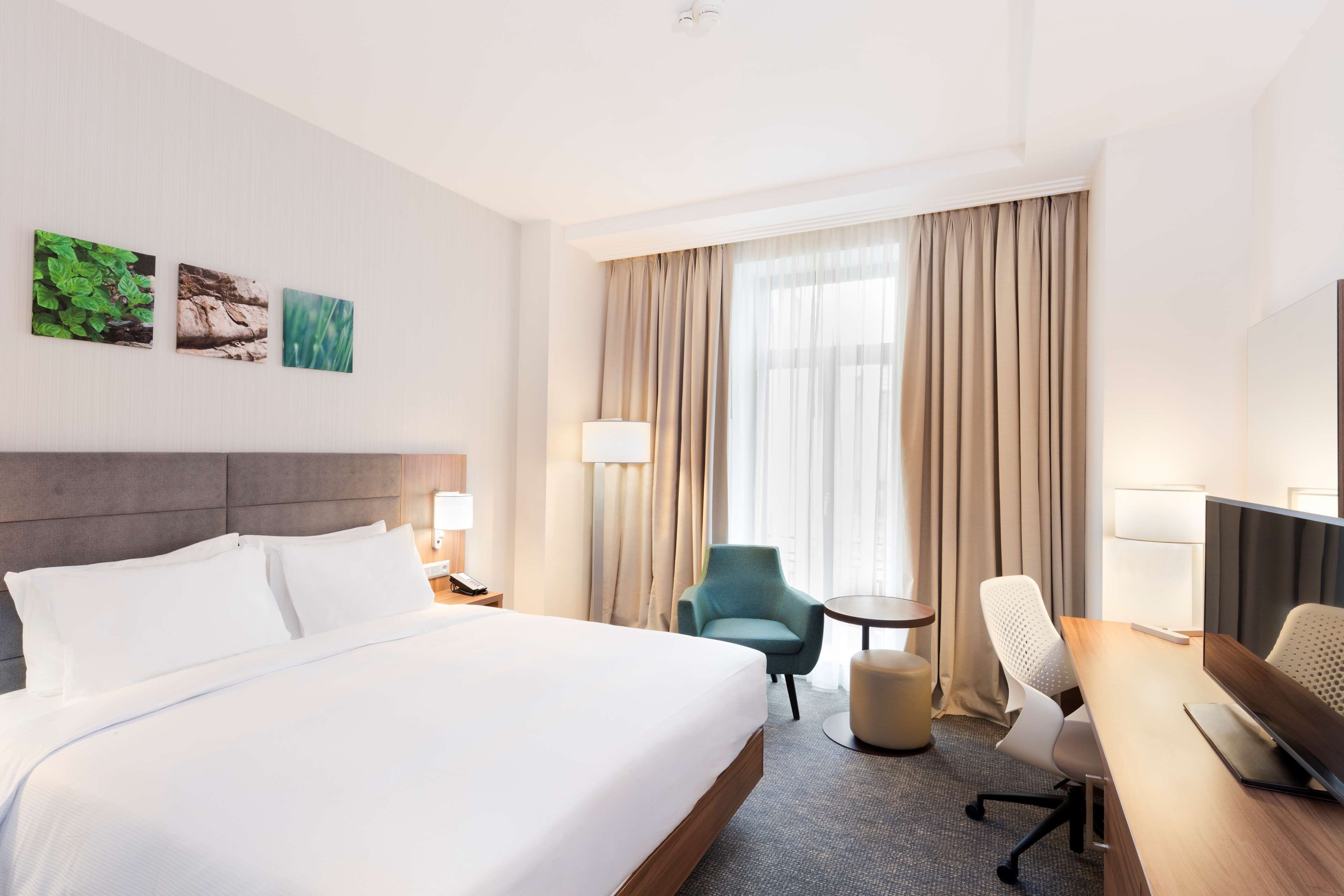 A hotel room with a large bed, a desk with a computer, a chair, a small table, and a window with curtains.