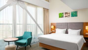 A hotel room with a large bed, a teal armchair, a small round table, and a view of the city through large windows.
