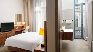 A modern hotel room with a large bed, a flat-screen TV, a bedside table with a lamp, a window with sheer curtains, and a bathroom with a sink and mirror.