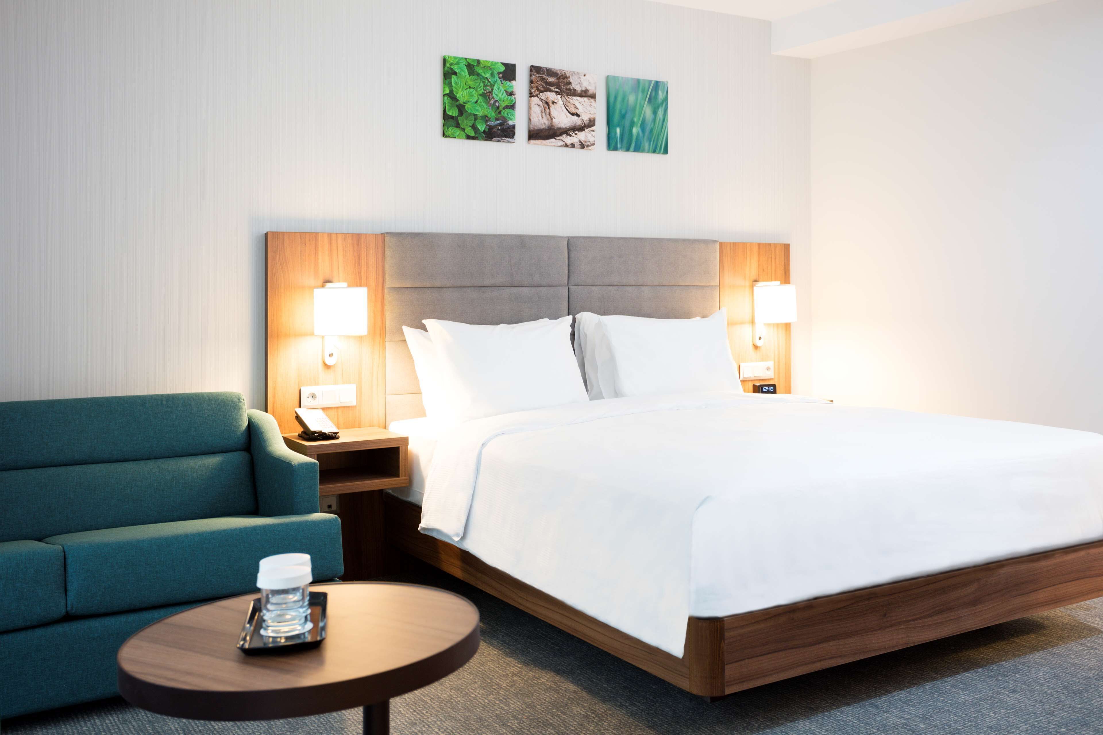 A hotel room with a bed, a green armchair, a bedside table with a lamp, and a small round table with a glass on it.