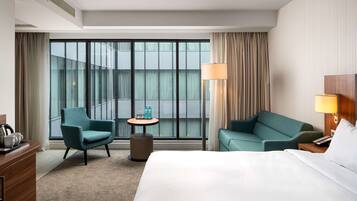 Habitación de hotel con una cama grande, dos sillones, una mesita redonda con una lámpara y vistas de la ciudad a través de amplias ventanas.