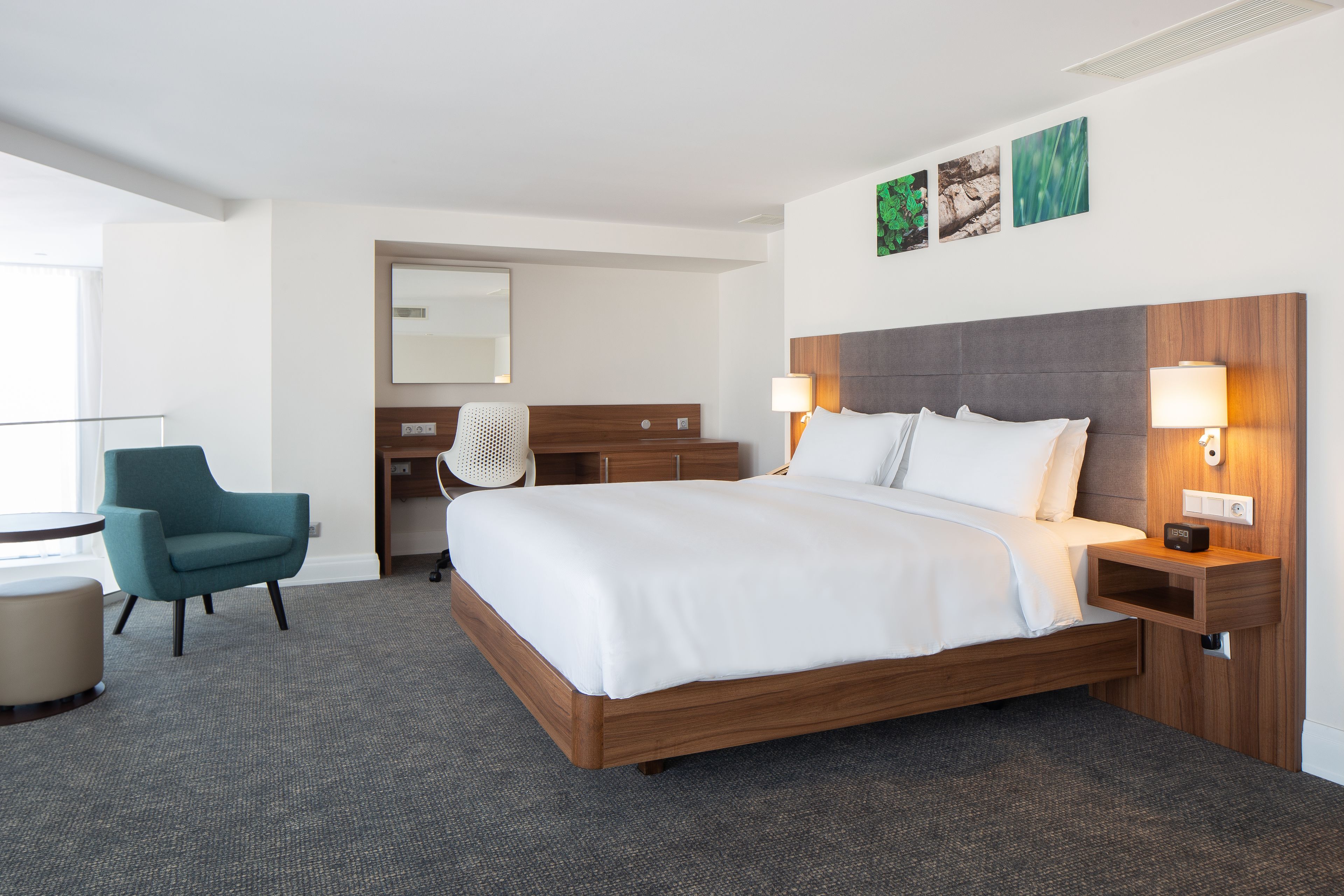 A hotel room with a large bed, two bedside tables with lamps, a desk with a chair, a mirror, and artwork on the wall.