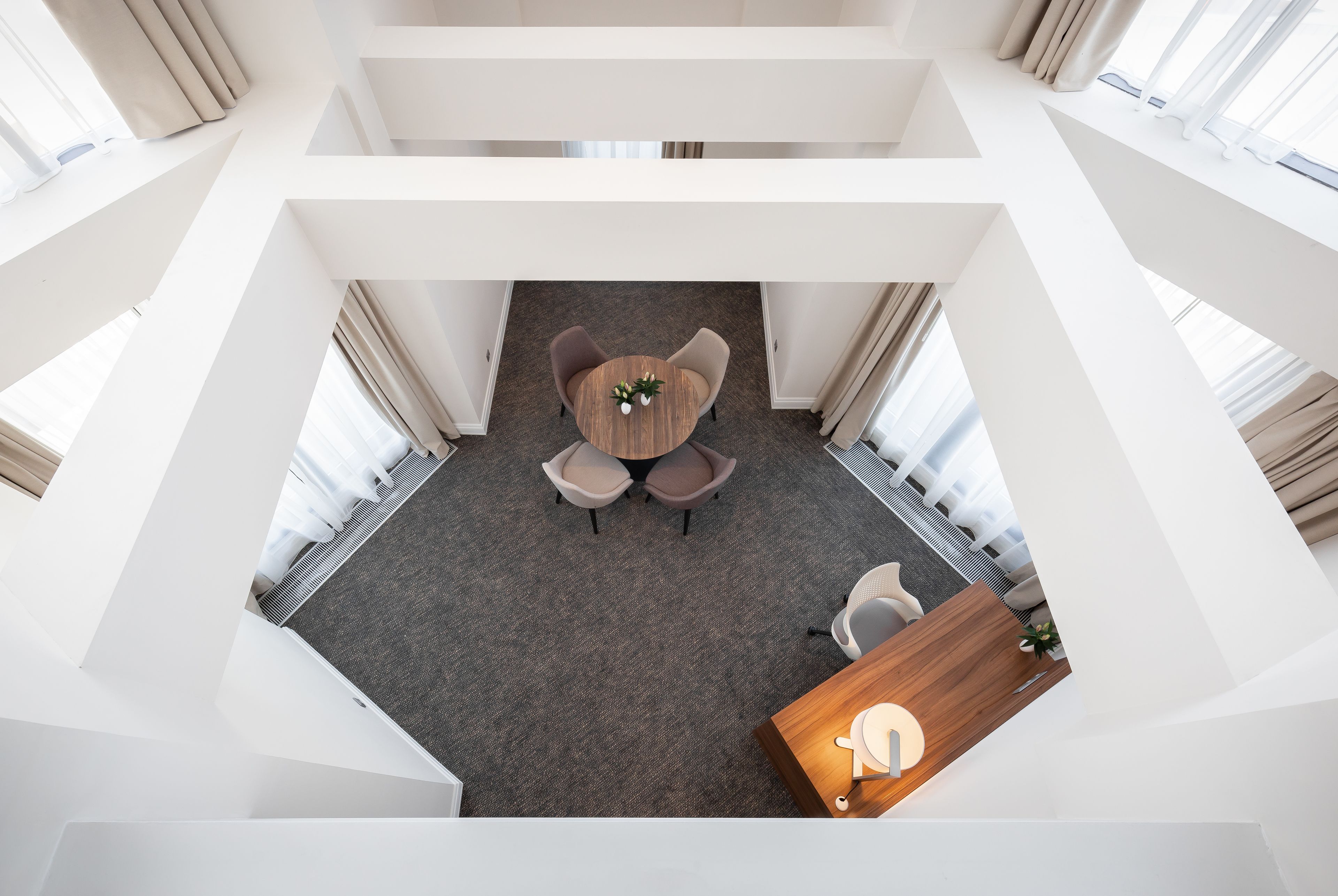 A modern hotel room with a central round table, two chairs, a small plant, and a wooden side table with a lamp.