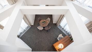 A modern hotel room with a central round table, two chairs, a small plant, and a wooden side table with a lamp.