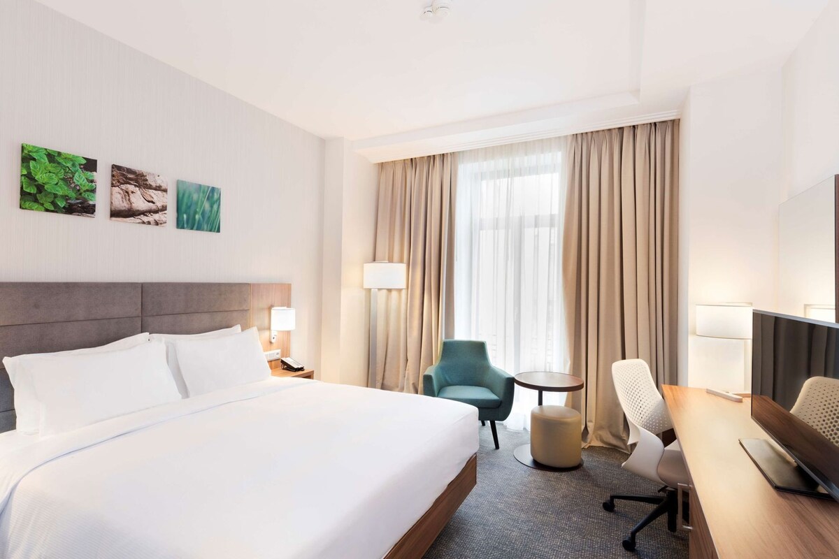 A hotel room with a large bed, a desk, two chairs, a lamp, and a window with curtains.