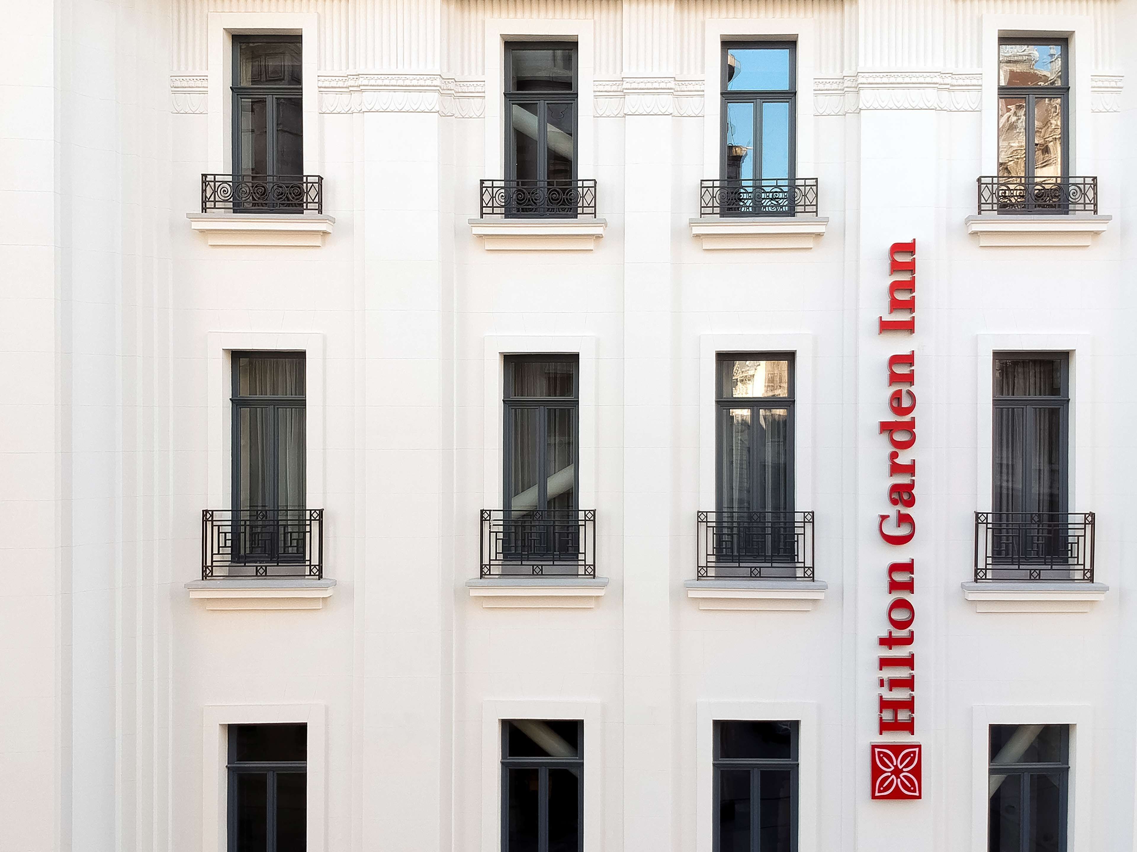 A multi-story building with a white facade and black window frames. The building has a series of balconies with black wrought iron railings. A large red sign with the text "Hilton Garden Inn" is prominently displayed on the right side of the building.