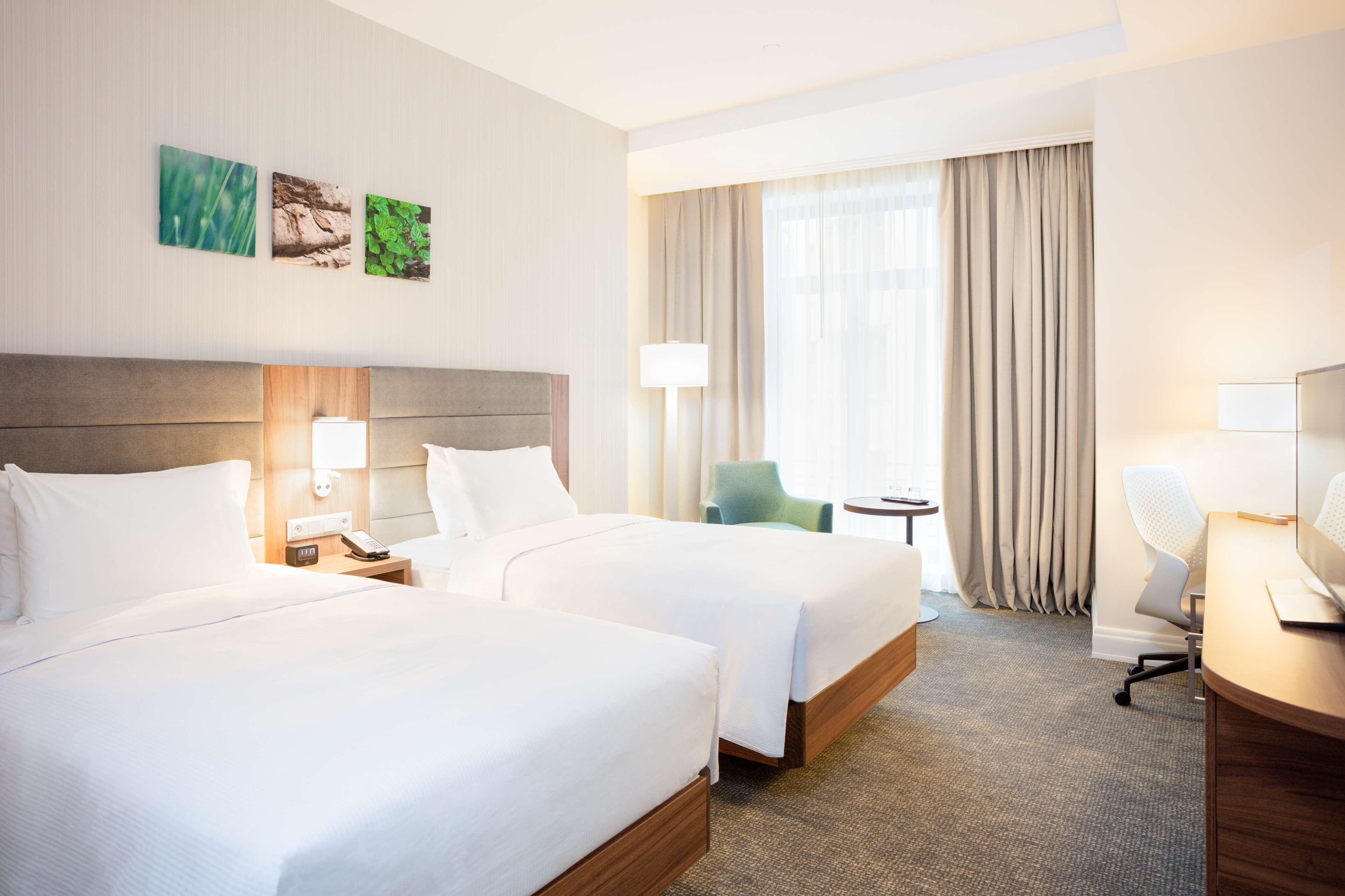 A hotel room with two beds, a desk, a chair, and a small table. There are three wall-mounted artworks and a large window with curtains.