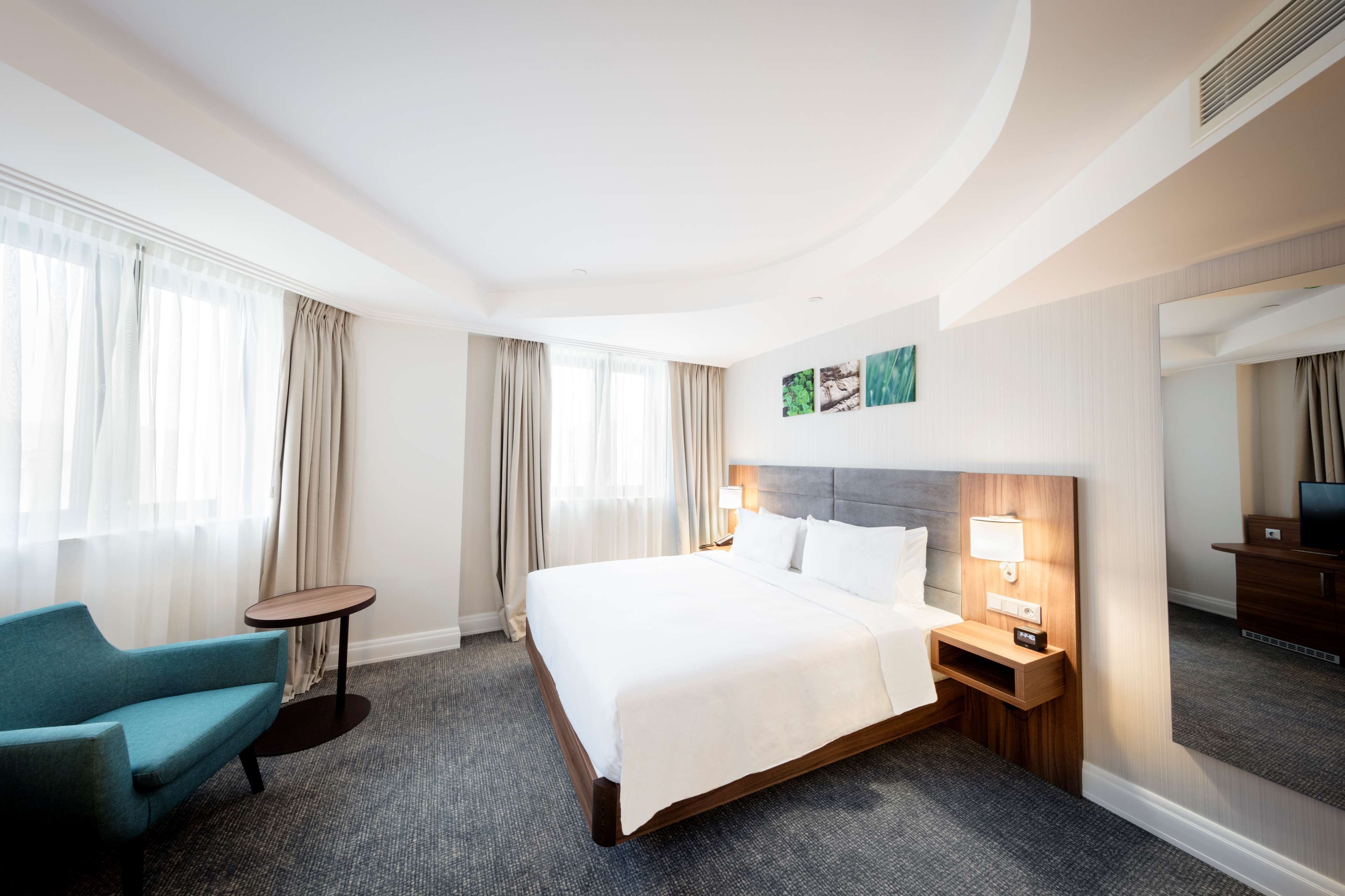 Una camera d'albergo con un letto grande, un comodino con una lampada, un tavolino rotondo, una poltrona blu e un grande specchio.