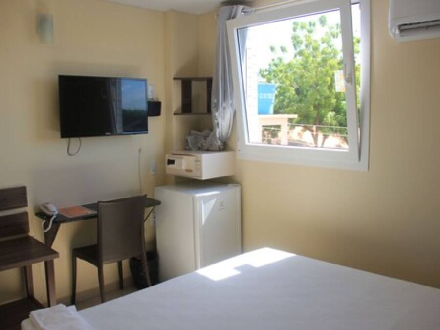 Habitación de hotel con cama, escritorio con silla, televisión, refrigerador pequeño y ventana con vista a árboles.