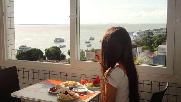 A dining area with a table set for breakfast, a window with a view of the sea and boats, and a person sitting at the table. - Sandis Mirante Hotel (Santarém)