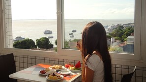 Un comedor con una mesa dispuesta para el desayuno, una ventana con vistas al mar y barcos, y una persona sentada a la mesa.
