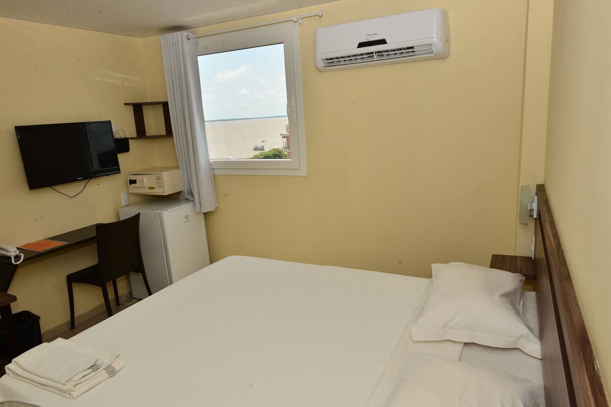 Habitación de hotel con cama, escritorio, silla, televisor, aire acondicionado y ventana con vista al mar.