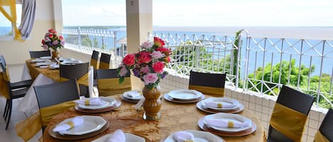 A dining table set for six with a view of the sea, a white railing, and a yellow and gold tablecloth.
