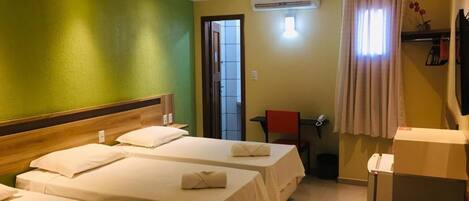 A hotel room with three single beds, a wooden headboard, a small table, a chair, an air conditioning unit, a curtain, and a television.