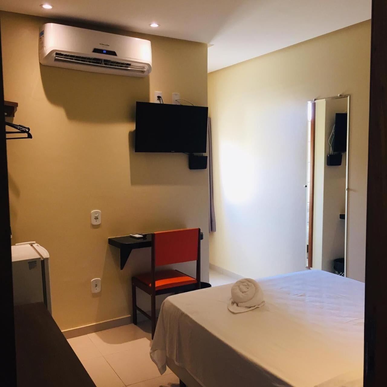 A hotel room with a bed, a chair, a TV mounted on the wall, an air conditioning unit, and a mirror.