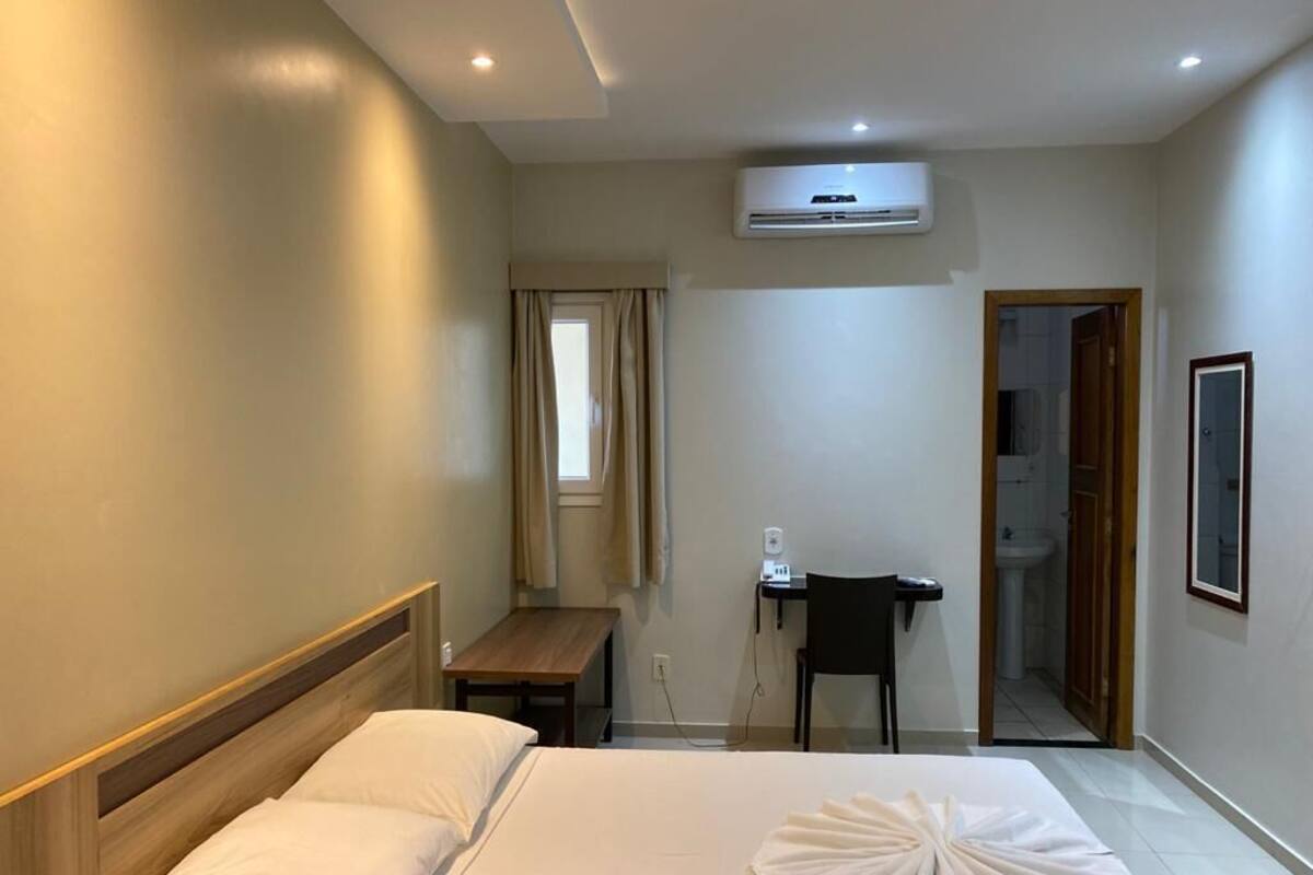A hotel room with a bed, a desk, a chair, an air conditioning unit, a window with curtains, and a door leading to a bathroom.