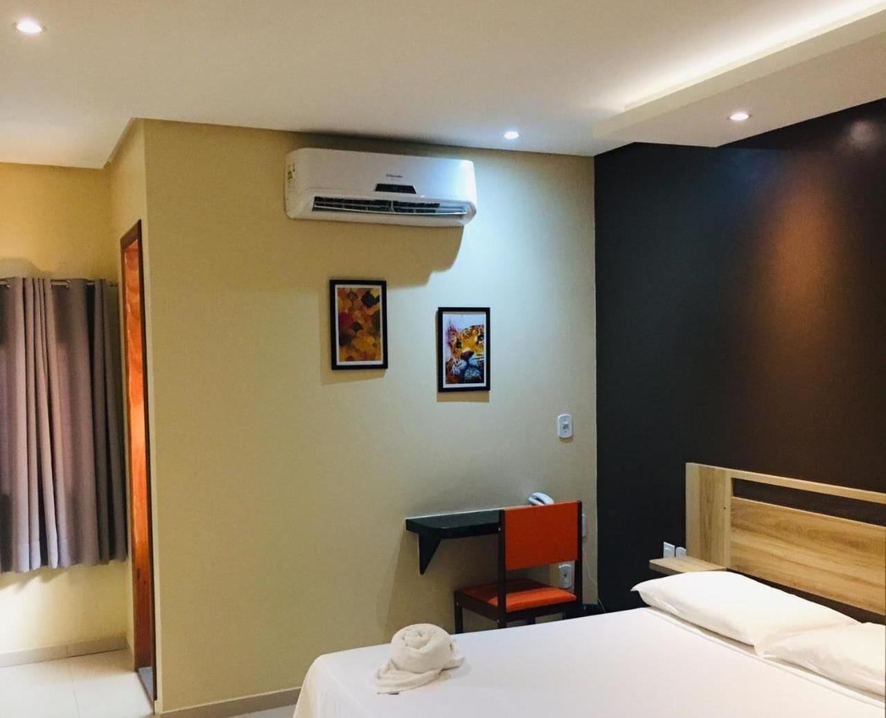 A hotel room with a bed, a desk, an air conditioner, and two framed pictures on the wall.