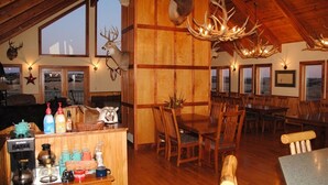 A rustic interior with wooden beams, a dining area with wooden chairs, a chandelier with antlers, and deer heads on the walls.