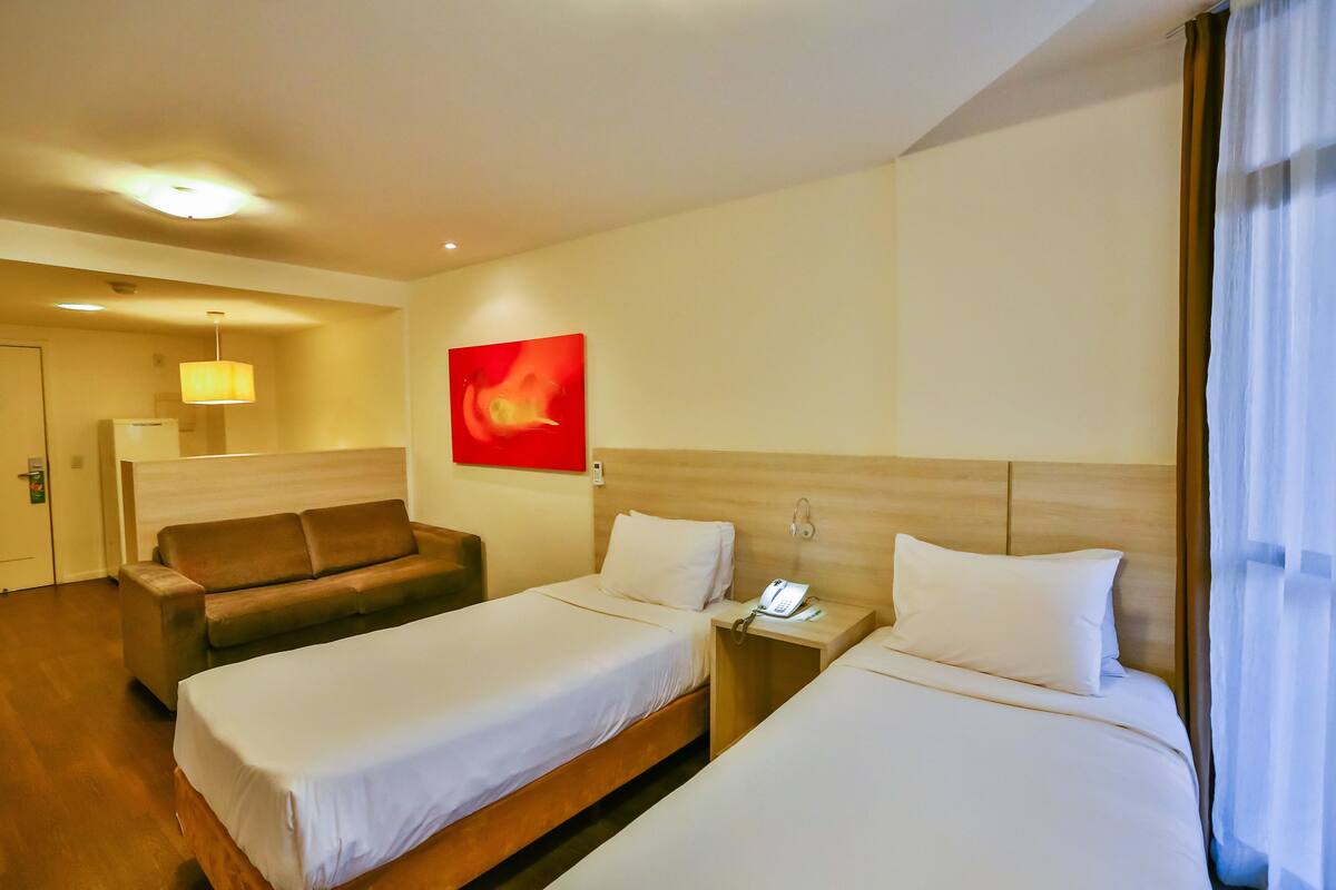 A hotel room with two beds, a sofa, a bedside table with a phone, and a red abstract painting on the wall.